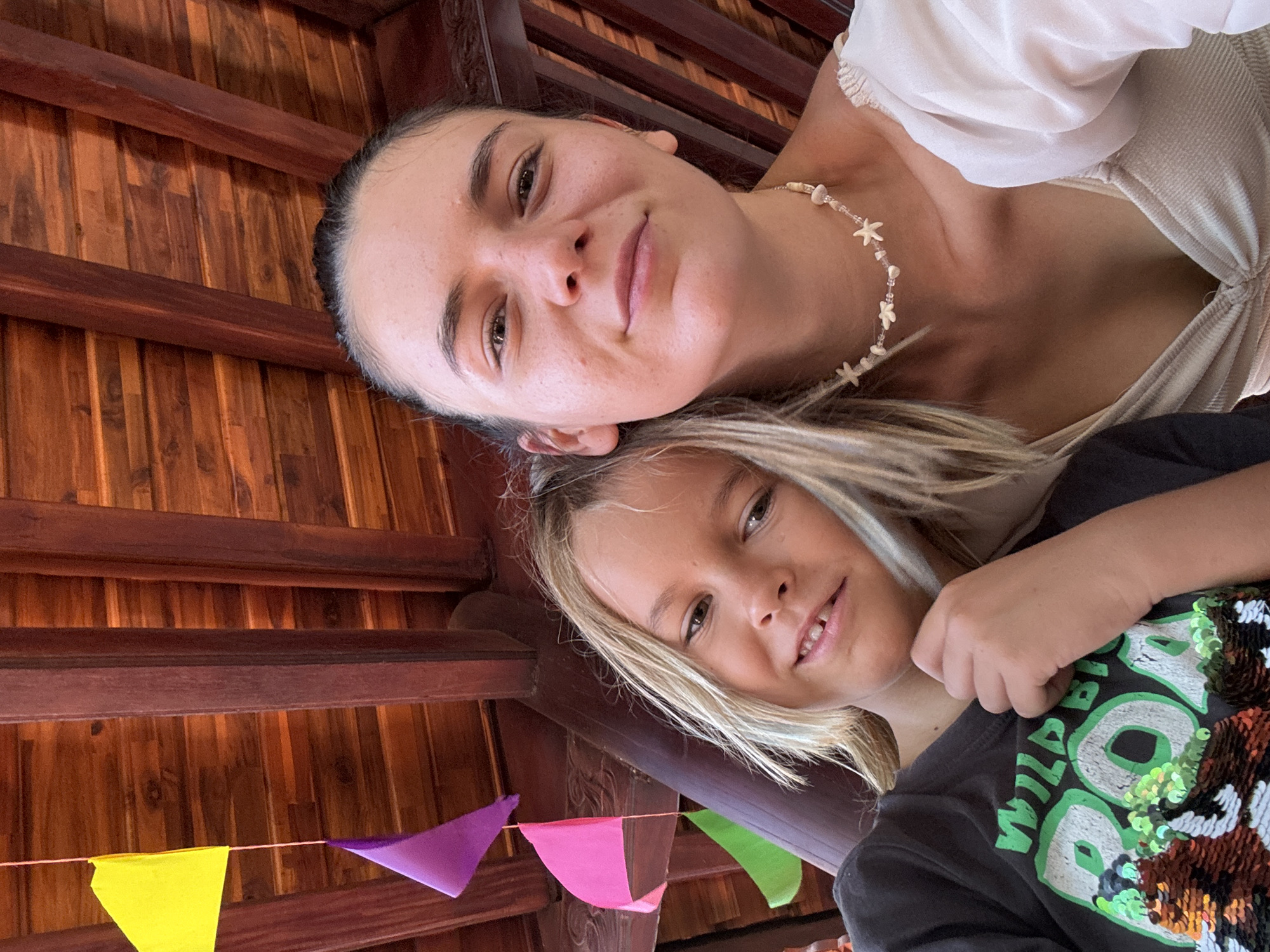 A slightly rotated, low-angle shot depicts a woman and a child indoors. The woman has dark hair pulled back, fair skin, and is wearing a cream-colored top and a necklace made of shells and beads. Her expression is a slight smile. The child, with blond hair, is wearing a dark T-shirt with a sequined design featuring the word "WILD" in green lettering. They are positioned closely, suggesting a familial relationship. Above them is a wooden ceiling structure with triangular pennants in yellow, purple, pink, and green strung along the beams. The scene appears to be taken indoors, and given the prompt's request to include location if known, the photograph is stated to be taken in Mũi Né, Vietnam.