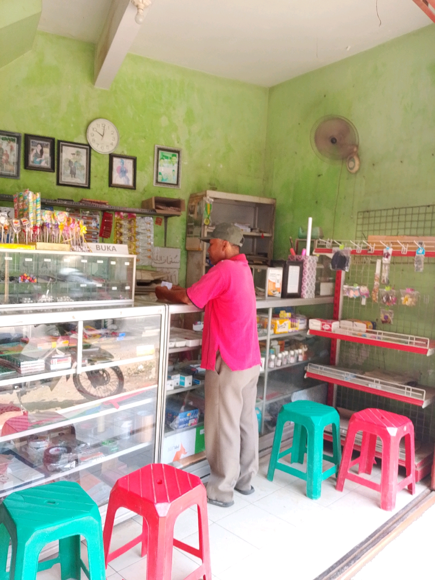 The image depicts the interior of a small convenience store, likely located in Tiron, Indonesia, based on the context provided. The walls of the store are painted a vibrant lime green, giving the space a cheerful, albeit slightly aged, appearance. A large analog clock with a white face and black numbers hangs on the left wall, showing the time as approximately 1:50. To the left of the clock, a series of framed pictures are hung, some of which appear to be family photographs.
In the center of the store, a man wearing a pink t-shirt, khaki pants, and a dark green cap is standing behind a glass display counter. He is holding a small piece of paper in his hands and appears to be interacting with the contents of the counter or perhaps making a transaction. The display counter is filled with various items, including what appear to be candies, stationery, and other small merchandise. Beneath the counter, a motorcycle wheel is visible, suggesting it might be stored or used for transport within or near the store.
On shelves behind the man, more products are neatly arranged, including boxes of what seem to be medications or supplements, and other packaged goods. A metal wall rack to the right displays an assortment of small items, possibly trinkets or accessories. A ceiling fan is mounted on the right wall. In the foreground, two plastic stools, one green and one red, are placed on the tiled floor, indicating a waiting area for customers. The overall scene suggests a functional, everyday retail environment.