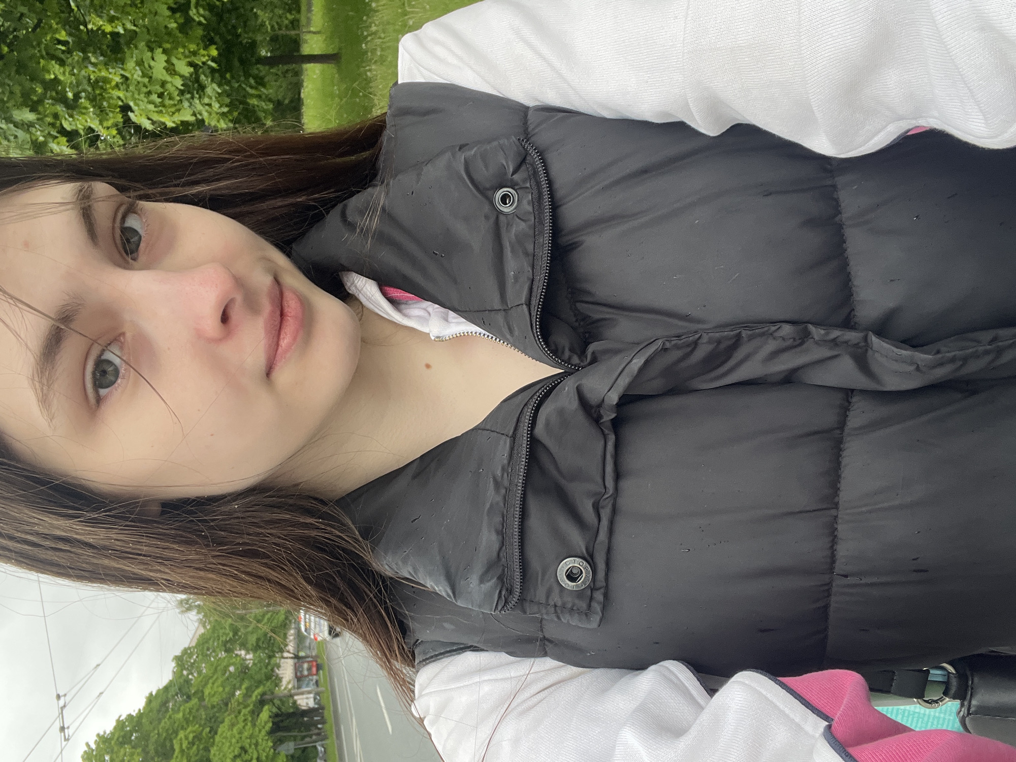 A young woman takes a selfie outdoors. She has light skin, light eyes, and dark, shoulder-length hair. She is wearing a white long-sleeved shirt and a black, unzipped puffer vest. The vest appears damp with water droplets.
In the blurred background, green trees and a street with cars are visible, suggesting an urban park or street setting. Based on architectural styles visible in the image, this photograph appears to be taken in Saint Petersburg, Russia.