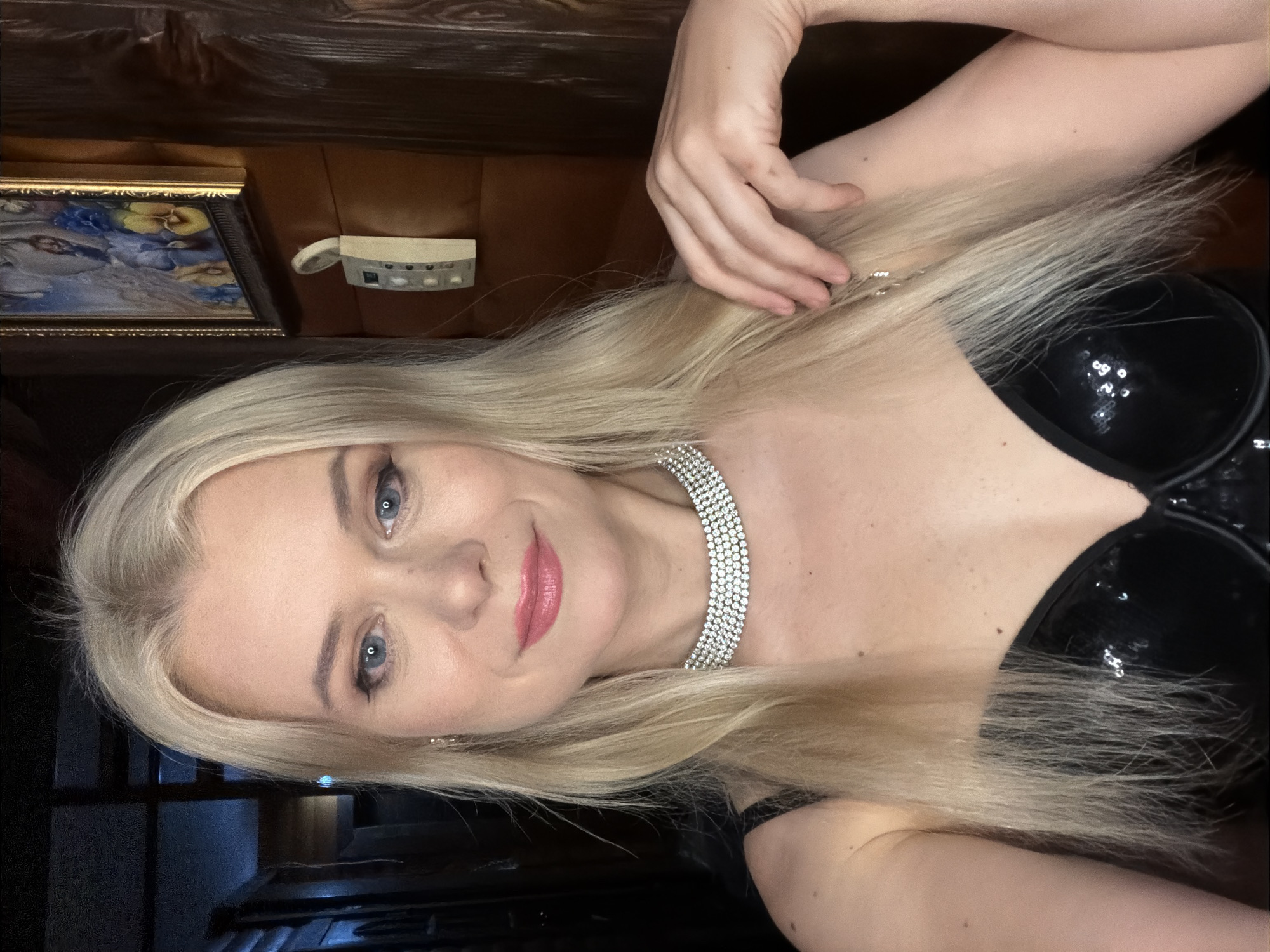 A blonde woman is taking a selfie in a dimly lit room with wooden walls. She is wearing a black sequined bustier and a diamond choker. Her left arm is bent, with her hand resting near her hair, and her right arm is out of frame. In the background, a framed painting and a wall-mounted intercom system are visible. The room's interior suggests a rustic or cabin-like setting. The image does not contain any visual elements that would indicate a specific location in Sochi, Russia.