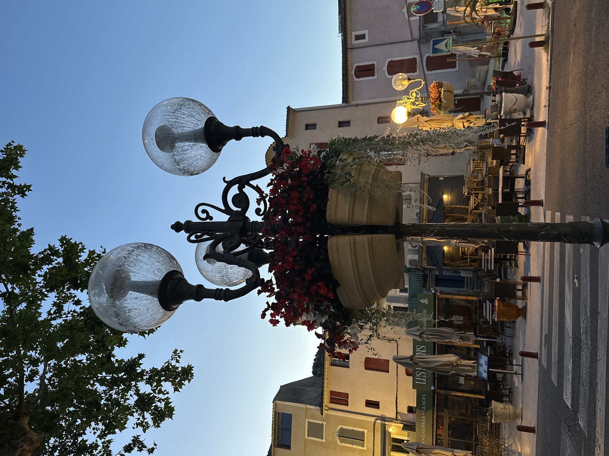 The image depicts a street scene in Anduze, France, at dusk. A lamppost with three glass globes and a planter filled with red and white flowers stands in the foreground. Behind the lamppost, buildings with closed shutters line a street where outdoor seating for restaurants is set up. A crosswalk is visible in the lower part of the image. The sky is a clear twilight blue.