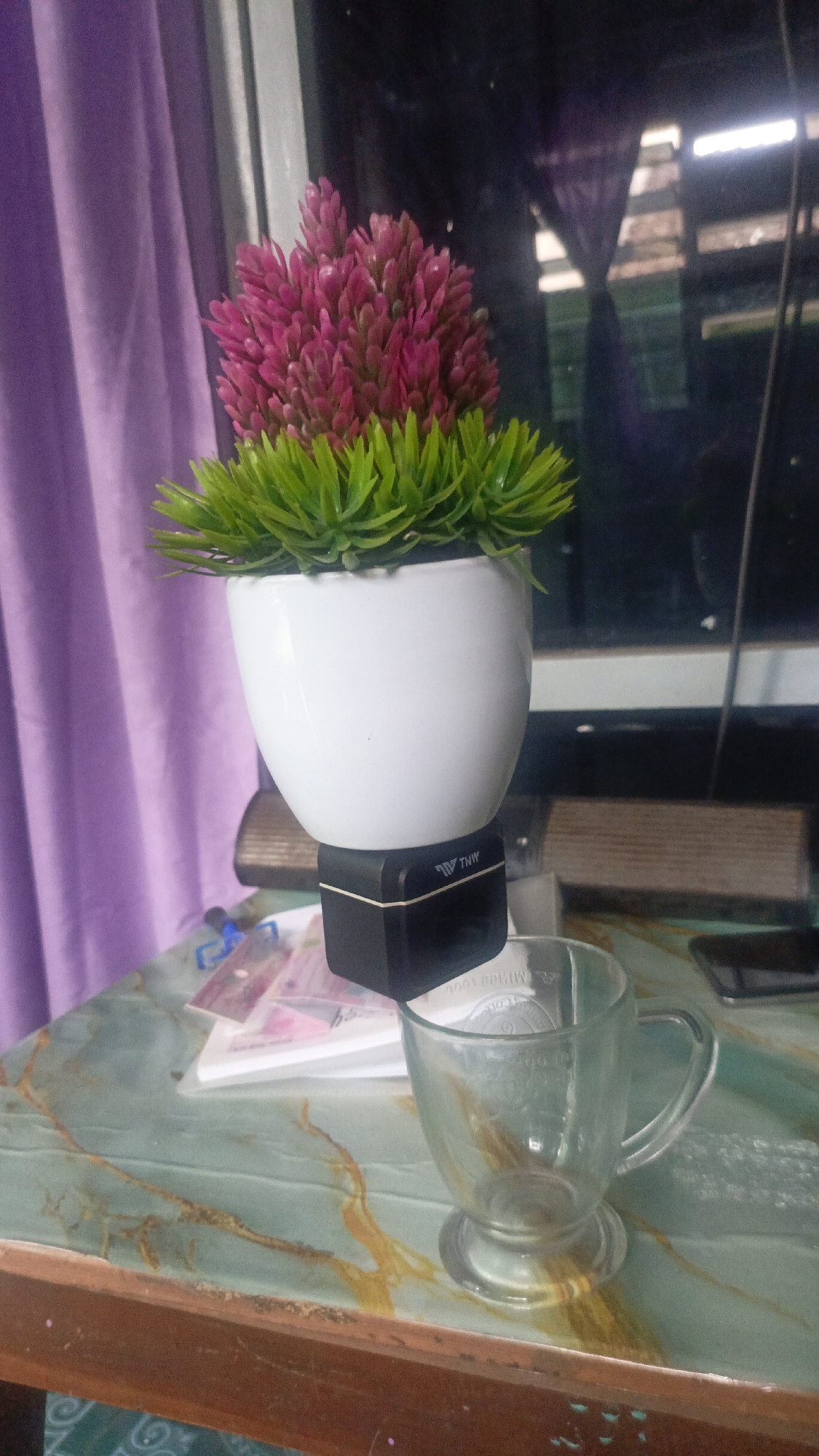 In Salatiga, Indonesia, an artificial pink and green plant sits in a white pot atop a black cube. To the right of the plant, an empty clear glass mug with a handle rests on a table. The table has a green and brown marble-like pattern. In the background, purple curtains are visible on the left, and a dark reflection of what appears to be a room with windows is on the right.