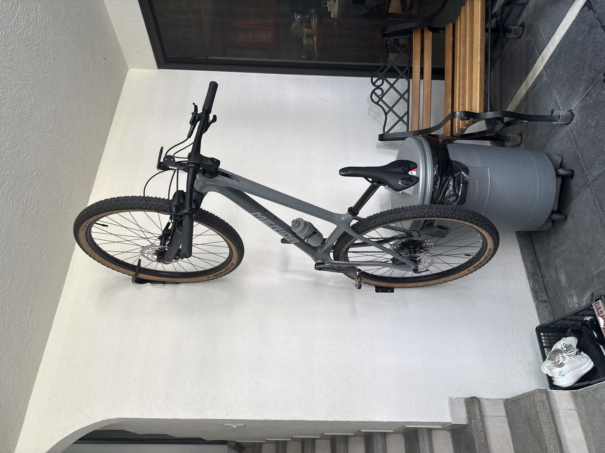A gray mountain bike is mounted horizontally on a white wall with a textured surface. The bike has a water bottle attached to its frame. Adjacent to the bike, a gray trash can stands upright, partially obscured by a black trash bag. Further to the right, a black plastic basket contains a pair of white sneakers and other items. The scene is located near a set of concrete steps, hinting at an indoor or semi-outdoor setting. A bench with black metal legs is behind the trash can. The image was taken in San Bernardino Tlaxcalancingo, Mexico.