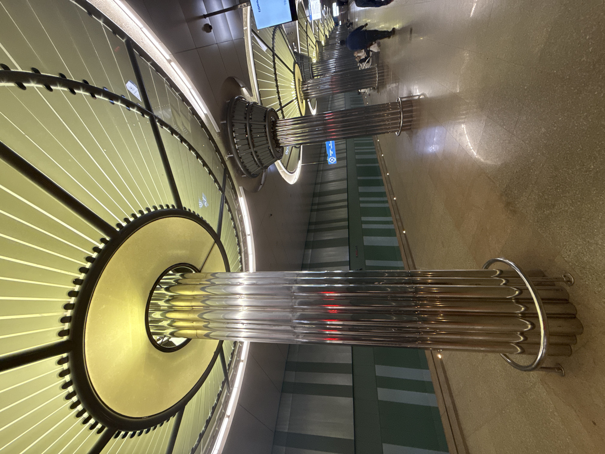 The image depicts the interior of a modern metro station, likely in Nizhny Novgorod, Russia, given the context provided. The foreground is dominated by a large, fluted chrome pillar supporting an elaborate, fan-shaped ceiling with a translucent yellow material illuminated from behind by white lights, creating a radiating pattern. The pillar has a metallic sheen and reflects red light from an unseen source.
The platform area is visible beyond the pillar, with a polished floor that reflects the overhead lighting. Further down the platform, more similar chrome pillars and illuminated ceiling structures are visible, receding into the distance. The station walls are lined with alternating green and pale vertical stripes.
Several individuals are present on the platform. In the middle ground, a person is walking away from the camera towards the right. To the right of this person, other individuals are seated on benches. In the background, a digital display screen is visible on the wall. The overall impression is one of contemporary design and spaciousness within the metro station.