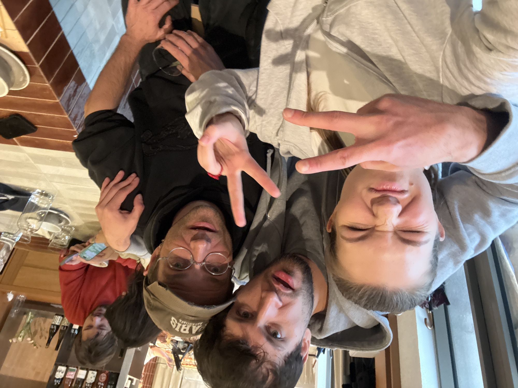 The image depicts an indoor scene with five individuals, likely in a casual dining establishment or bar. The specific location within Belgrade, Serbia, cannot be determined from the visual information.
In the immediate foreground, three subjects are posing for a close-up photograph.
1. On the left, a young adult female with hair pulled back wears a light grey zippered hoodie over a white top. She exhibits a humorous expression, with eyes squeezed shut and lips pursed, and displays a "peace" sign with her left hand.
2. In the center, a young adult male with dark hair and stubble wears a grey hooded sweatshirt. His left arm is around the female's shoulders. He looks directly at the camera with an open-mouthed expression.
3. On the right, an adult male with dark hair, wearing circular wire-rimmed glasses and a distressed olive-green baseball cap inscribed "PARIS Balenciaga," wears a dark hooded sweatshirt. His right arm is around the central male's shoulders, and he displays a "peace" sign with his right hand. He holds a clear glass containing liquid in his left hand and looks at the camera with an open-mouthed expression.
In the mid-ground, two additional individuals are seated at a table behind the foreground group.
4. To the far right, an adult male with dark hair, wearing a red long-sleeved shirt, is engrossed in a smartphone held in his hands.
5. Partially obscured behind this male, another individual with dark hair leans forward, seemingly observing the smartphone.
The environment includes a table with a patterned surface (alternating brown and lighter tiles) holding several clear glasses, white cups, saucers, and a clear bottle. A dark wooden shelf unit behind the table displays multiple bottles, likely alcoholic beverages, and a small vase with flowers. A folded dark umbrella hangs on a hook in the far left background. The scene is brightly lit.