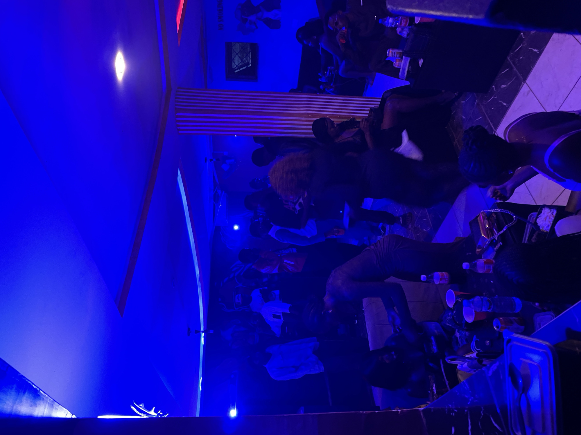 This is a nighttime, indoor photograph taken in what appears to be a dimly lit lounge or club in Wumat, Nigeria. The scene is dominated by vibrant blue and red lighting, casting a moody ambiance. Several individuals, predominantly young adults, are present and engaged in various activities.
In the foreground, a person in a dark top is bent over a table filled with drinks and other items. To the left, a counter area displays containers and utensils, suggesting food or beverage service.
The midground features a group of people gathered around couches and seating areas. Some are socializing, while others appear to be resting or observing the surroundings. A prominent striped column is visible in the center of the room.
In the background, more individuals are seen, some standing and others seated. Artwork or murals adorn the walls, with a "NO SMOKING" sign also visible. The ceiling is illuminated with strip lighting and a central fixture. The overall impression is that of a social gathering at an establishment that offers entertainment and refreshments.