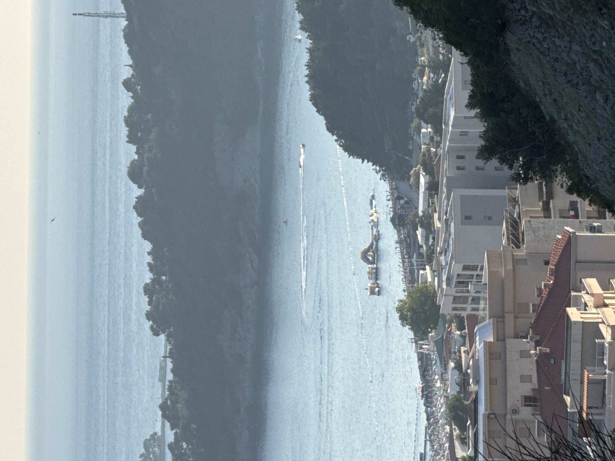 This is a distant, high-angle, daytime, outdoor shot of the city of Rafailovići, Montenegro, capturing a section of the coastline and its surrounding landscape. The scene is dominated by the expansive, calm blue sea in the background, with a hazy horizon where the sky meets the water. A narrow strip of land covered in dense, dark green trees separates the sea from the inhabited area.
In the foreground, multiple buildings with light-colored facades and red-tiled roofs are visible. These appear to be multi-story residential or hotel buildings, some with balconies and visible windows. The architecture suggests a Mediterranean style. The buildings are nestled on a sloping hillside, with rocky terrain and scattered trees visible between them and in the lower right corner of the frame.
Further back, closer to the sea, is a section of a beach with numerous people, indicated by small dots, and umbrellas. Adjacent to the beach, in the water, there is a floating inflatable water park with various structures, including slides and platforms. Several small boats are visible in the water, with one motorboat creating a white wake. A series of buoys marks a boundary in the sea.
In the distance, on the tree-covered hills, a tall, slender telecommunications tower is visible on the right side. The overall atmosphere is peaceful and sunny, indicative of a coastal resort town. The image quality is somewhat soft due to the distance and possibly atmospheric conditions.