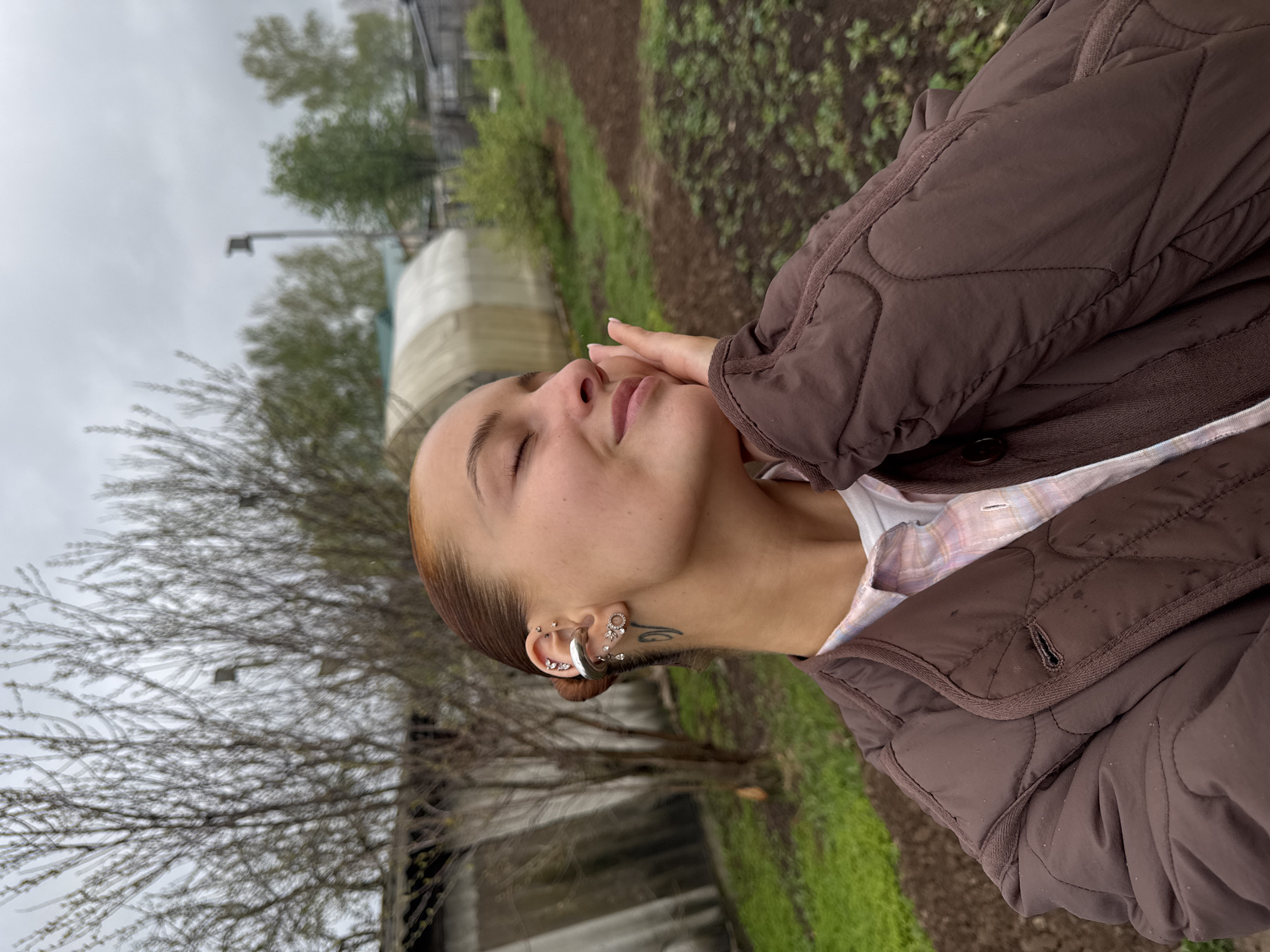 A medium shot captures a woman with light skin and brown hair, wearing a brown quilted jacket over a white and pink plaid shirt. Her eyes are closed, and her right hand is gently touching her cheek. She is adorned with multiple piercings in her left ear.
The background suggests a natural or agricultural setting, possibly in Chastyye, Russia. There is greenery along a path or row, suggesting cultivated land or a garden. A blurry structure, possibly a greenhouse or covered storage area, is visible in the distance. A bare tree stands to the left. The sky is overcast.