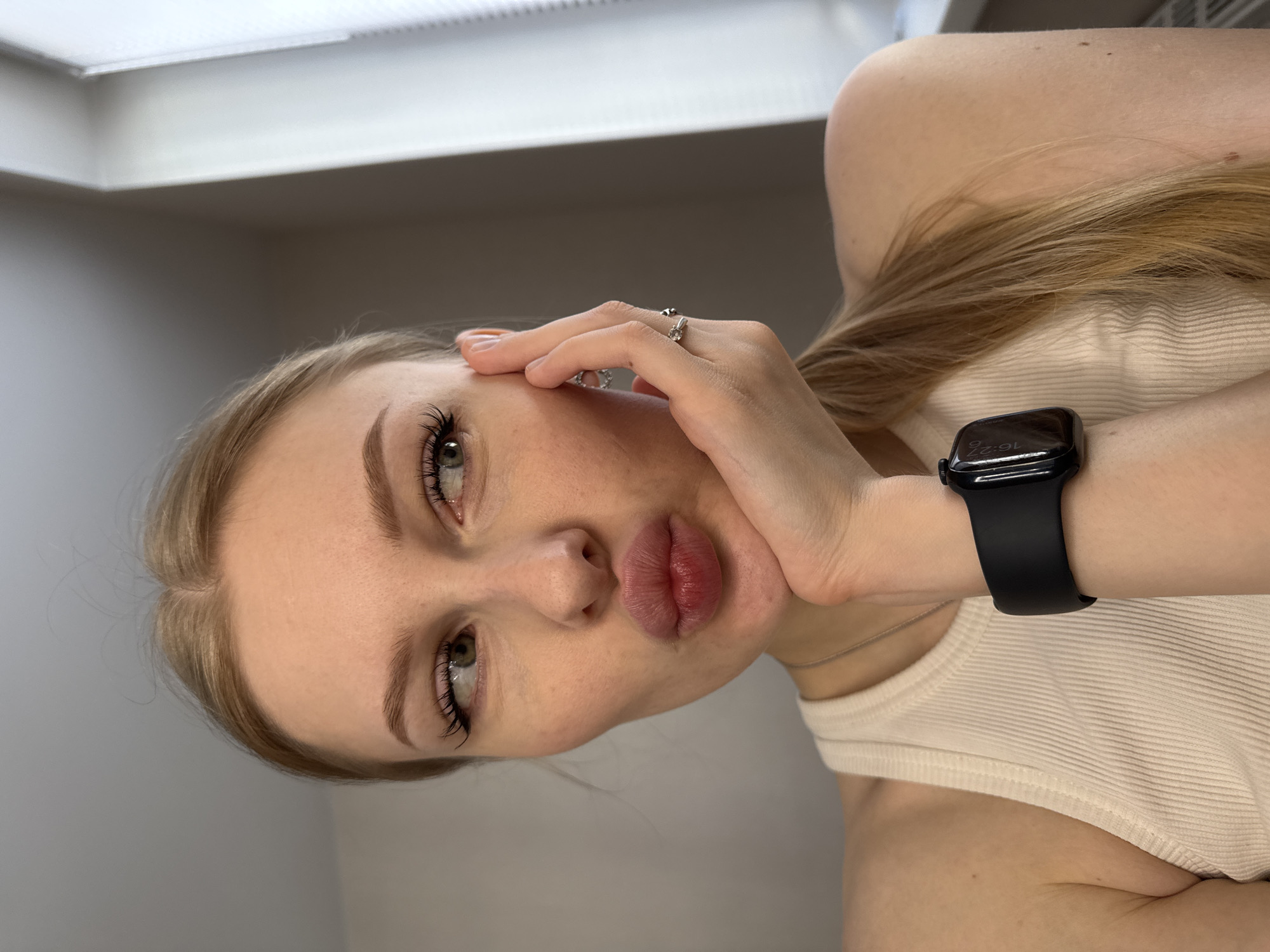 A blonde woman is shown in a medium close-up selfie. She wears a ring on her left ring finger and a black smartwatch on her left wrist. She has neutral makeup. Her right hand is touching her face. She is wearing a white tank top. The background is a gray wall and a white ceiling. The image appears to have been taken indoors. There is no information to confirm the image was taken in Perm, Russia.