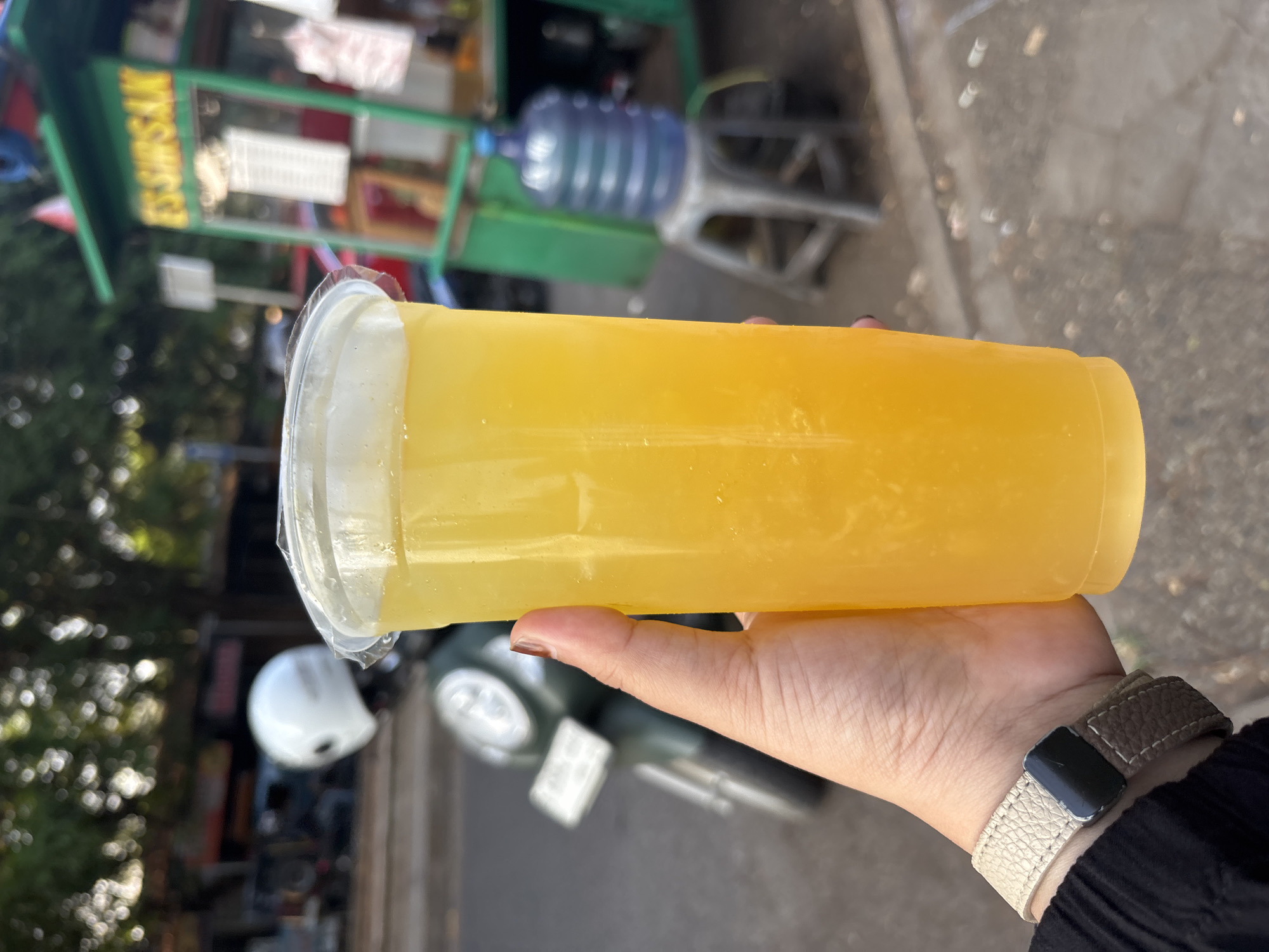 A close-up shot shows a hand holding a plastic cup filled with a bright yellow, translucent beverage, likely a fruit juice. The hand has a light skin tone and is wearing a beige and brown watch band. The beverage itself has some small bubbles and appears to be slightly pulpy. In the background, a green food cart with "ES SIRSAK" written on it in yellow letters is visible, suggesting it's a soursop juice vendor. A large blue water cooler bottle sits on a stool near the cart. Further back, a white helmet is placed on a dark-colored scooter, parked on an asphalt road. The environment appears to be an outdoor urban setting, possibly a street or market area in Cirebon, Indonesia, indicated by the presence of the food cart and local transportation. The overall lighting suggests it is daytime, with sunlight filtering through trees, creating blurred bokeh effects in the background.