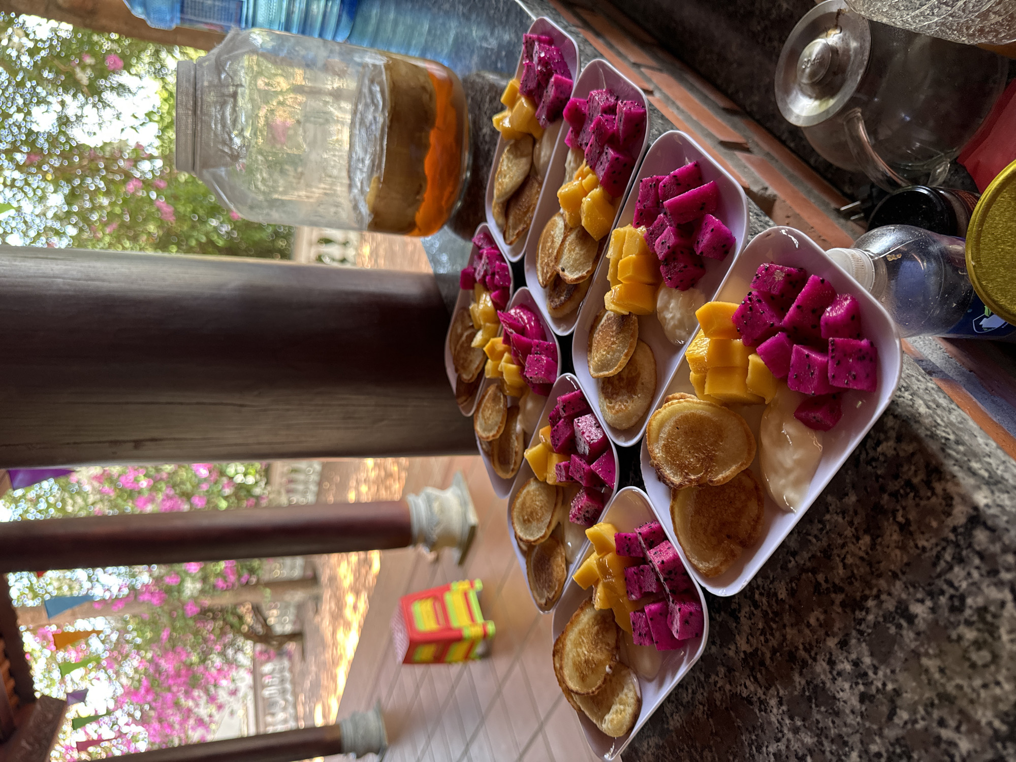 This media file captures an outdoor dining scene, likely a breakfast or brunch setup at a resort or guesthouse in Mũi Né, Vietnam. The image is taken from a slightly elevated perspective, looking down at a counter filled with numerous individual servings of food.
The primary subjects are rows of identical white rectangular dishes, each containing several small, round, golden-brown pancakes. Alongside the pancakes, each dish features a colorful assortment of diced fresh fruit: vibrant pink dragon fruit with visible black seeds, and bright yellow mango cubes. A dollop of a creamy white sauce, possibly yogurt or a sweet condiment, is also present on most plates.
In the background, a large clear plastic dispenser or jar with a distinct light brown/orange sediment or liquid at its base is visible, suggesting a flavored drink or juice. To the right, a glass teapot with a metal lid, a small clear plastic bottle, and a yellow-lidded container are also placed on the counter. The countertop itself appears to be dark granite or marble.
The setting is outdoors, framed by dark wooden beams or pillars. Beyond the immediate dining area, the background is softly blurred (bokeh effect), revealing lush green foliage interspersed with abundant bright pink or purple flowers, strongly resembling bougainvillea. Some colorful triangular flags or bunting are also visible in the distance, adding a festive touch.
The scene suggests a relaxed daytime meal, likely in the morning or early afternoon, given the bright natural light. The vibrant, tropical fruits and the outdoor, floral setting strongly align with the provided location context of Mũi Né, Vietnam, indicating a pleasant, resort-like atmosphere. No discernible text is visible in the image.