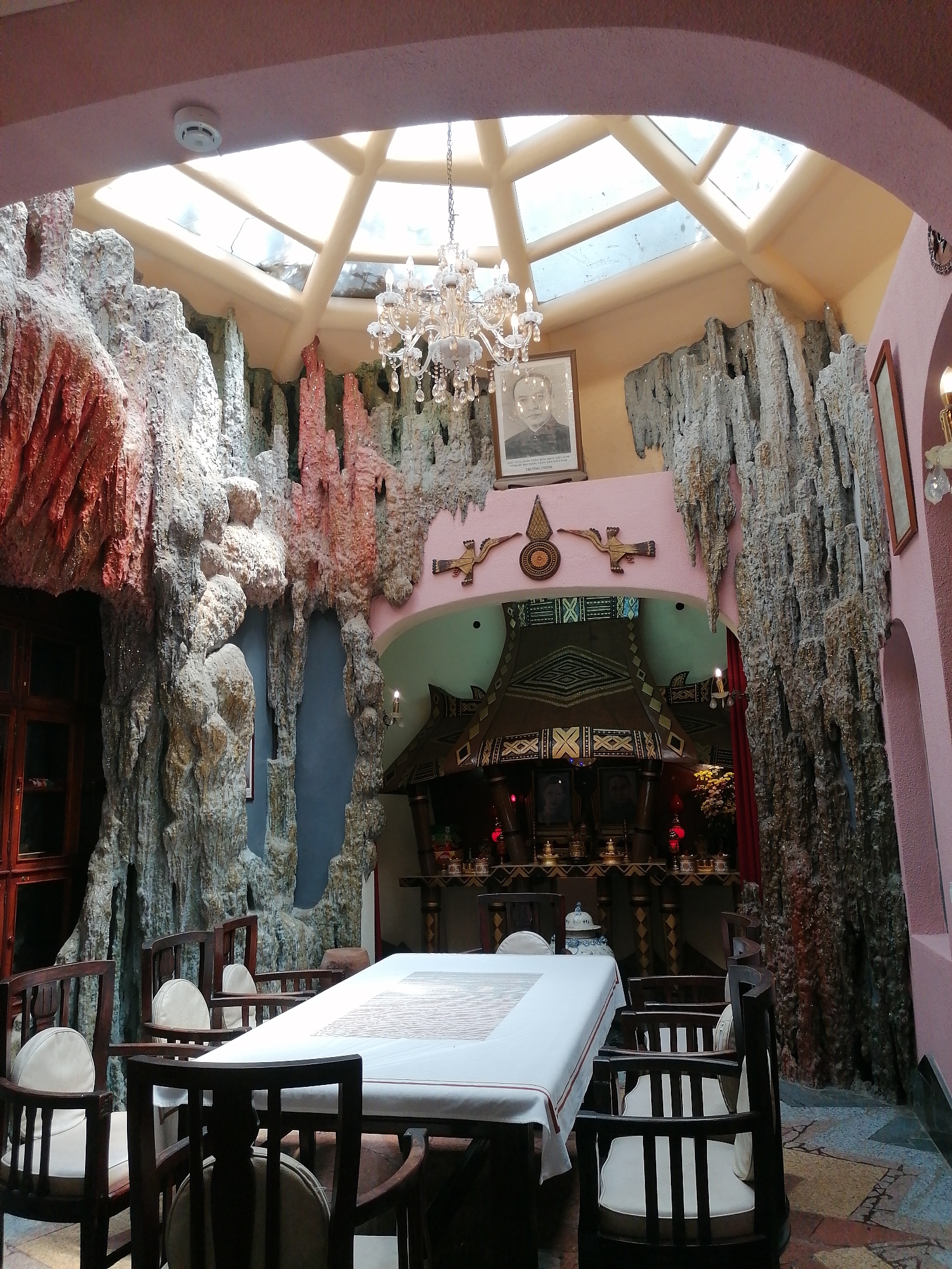 The image depicts the interior of the Crazy House in Da Lat, Vietnam. A rectangular table covered with a white tablecloth is surrounded by dark wooden chairs with white cushions. The walls mimic a cave environment with rocky textures and stalactite-like formations, painted in shades of grey, white, and red. An opening leads to a room with an altar-like structure decorated with intricate designs and lit by red candles. Above, a portrait hangs beneath a chandelier and a skylight that allows natural light to illuminate the room. The color palette includes pink walls, and the floor is patterned with tiles.