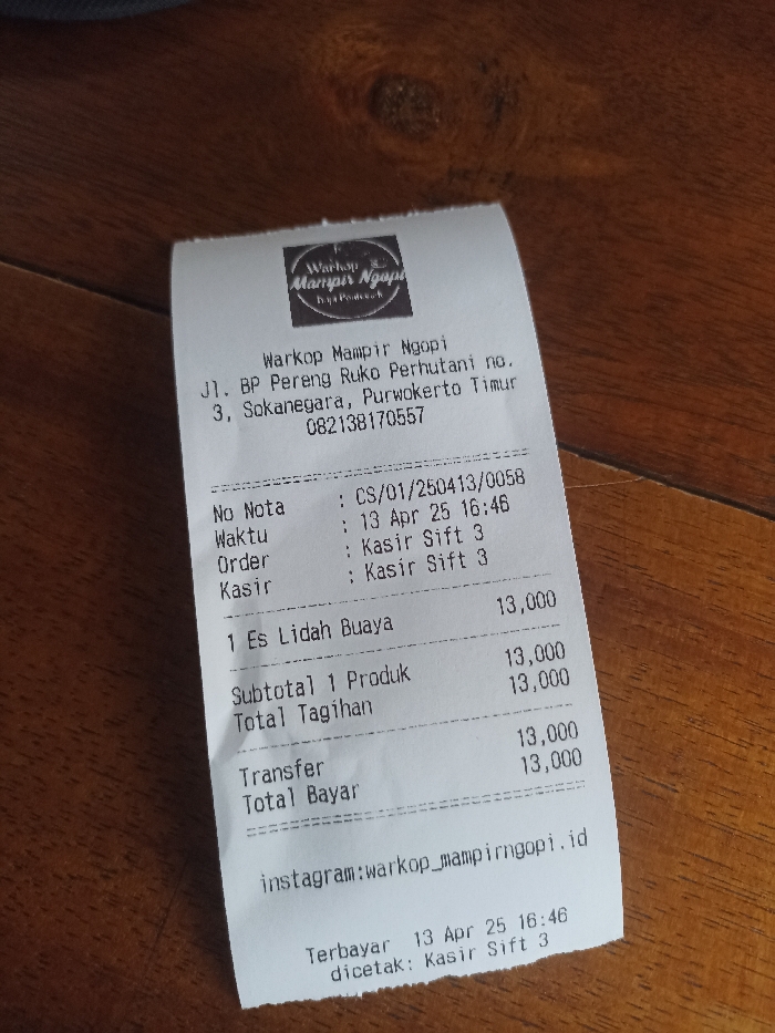 The image shows a receipt from a food stall called "Warkop Mampir Ngopi" located at Jl. BP Pereng Ruko Perhutani no. 3, Sokanegara, Purwokerto Timur, Indonesia. The phone number of the stall is 082138170557. The receipt was issued on April 13th, 2023 at 16:46, with order number CS/01/250413/0058, and was served by cashier shift 3. The receipt lists one item "1 Es Lidah Buaya" for 13,000 Indonesian Rupiah. The total bill was 13,000 Indonesian Rupiah, and the customer paid with a transfer. The receipt has an Instagram handle for the stall: "warkop_mampirngopi". The receipt was printed at 16:46 by cashier shift 3. The image is likely taken in a food stall or restaurant. The receipt is lying on a wooden surface.