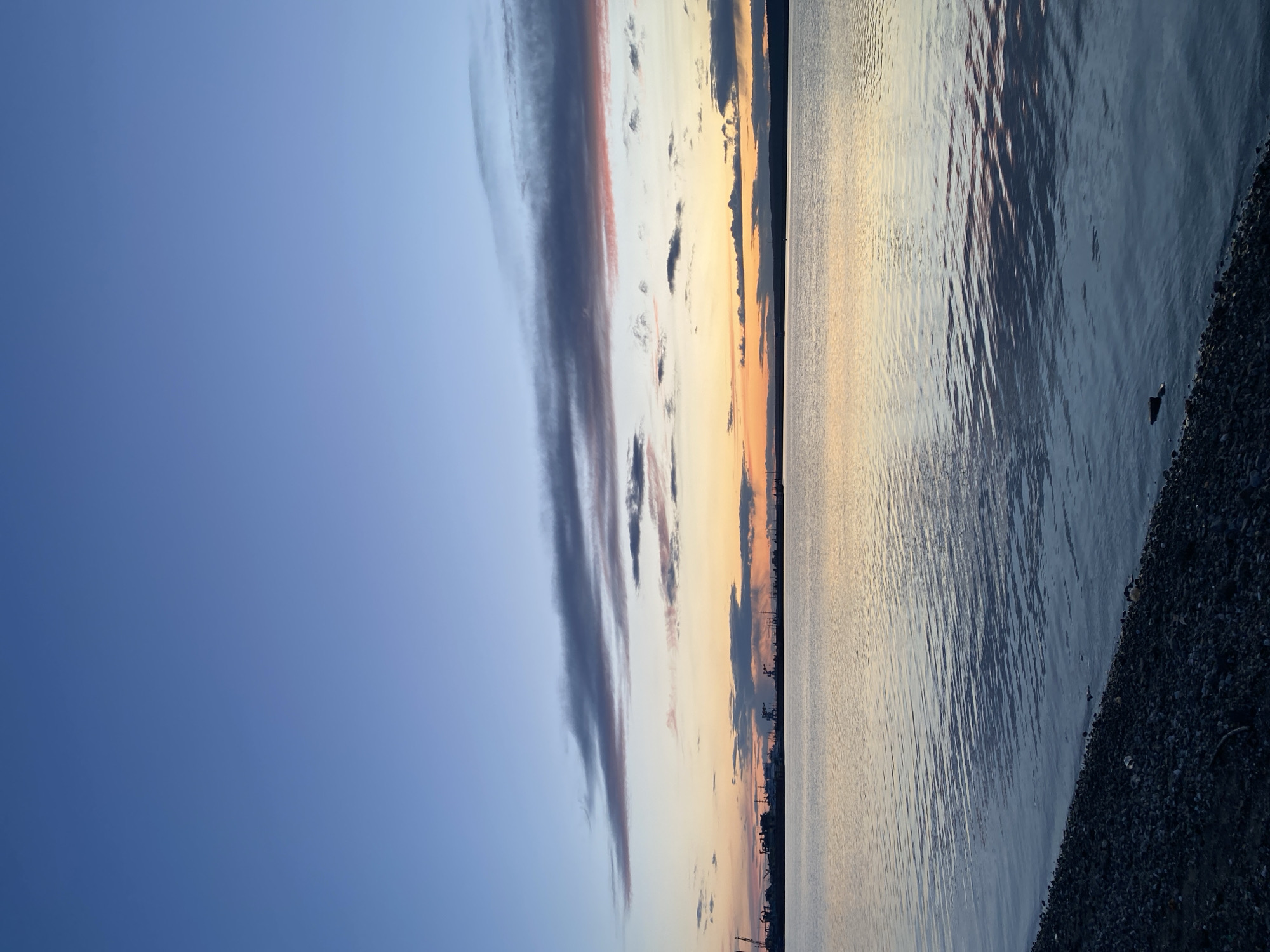 The image captures a tranquil waterscape at either sunrise or sunset, likely over the Kama River or Votkinsk Reservoir in Chaikovsky, Russia. The scene is dominated by a clear horizon line, dividing the sky from the water.
The sky, filling the upper portion, transitions from a soft, clear blue-grey on the left to warm shades of pale orange and yellow towards the right, indicative of twilight. Streaky, elongated clouds, ranging from dark grey to faint pink and orange, stretch across the sky, adding visual interest.
Below the horizon, the calm water reflects the colors of the sky, shimmering with light on the left and deepening into darker blues and grays on the right. Gentle ripples are visible on the water's surface, creating subtle patterns. Along the distant horizon, a faint, dark silhouette of what appears to be a distant cityscape or industrial structures is discernible, particularly towards the right side.
In the immediate foreground on the right, a dark, pebbly or rocky bank slopes gently into the water, adding texture to the scene. No individuals or specific activities are depicted, underscoring the peaceful and expansive nature of the landscape. The overall mood is serene and contemplative. There is no visible text in the media.