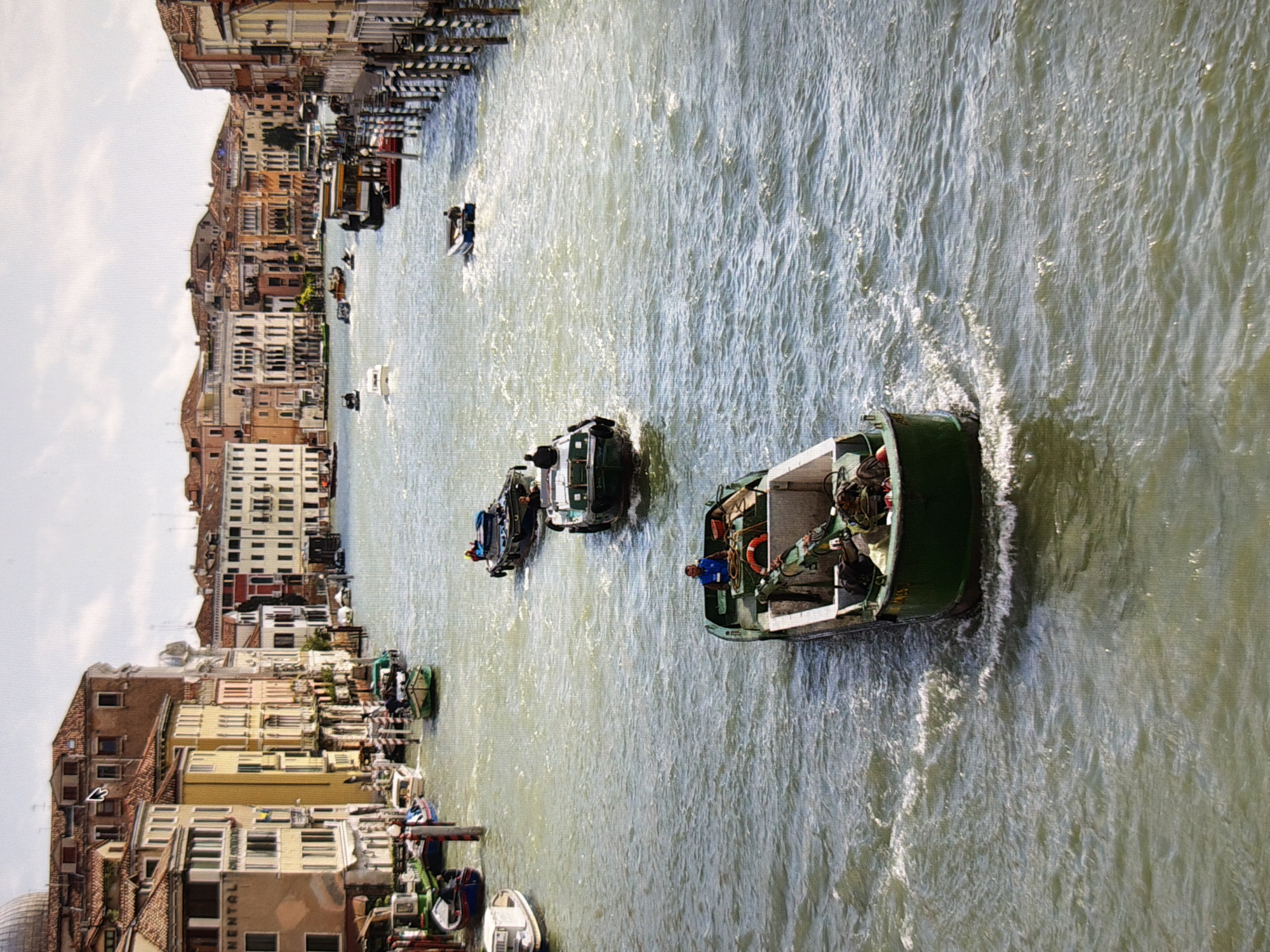 The image shows a wide canal or river with multiple boats navigating the water. Buildings with red tile roofs and stucco facades line the banks of the canal. On the right side, there are poles with black and white stripes. Several boats are visible, including a larger green boat in the foreground with a person in a blue shirt steering. Other boats are further down the canal, some carrying people. The water is choppy, creating ripples and whitecaps. The sky is partly cloudy. The scene appears to be in Venice, Italy, given the architecture and the canal system, not Kyiv, Ukraine.