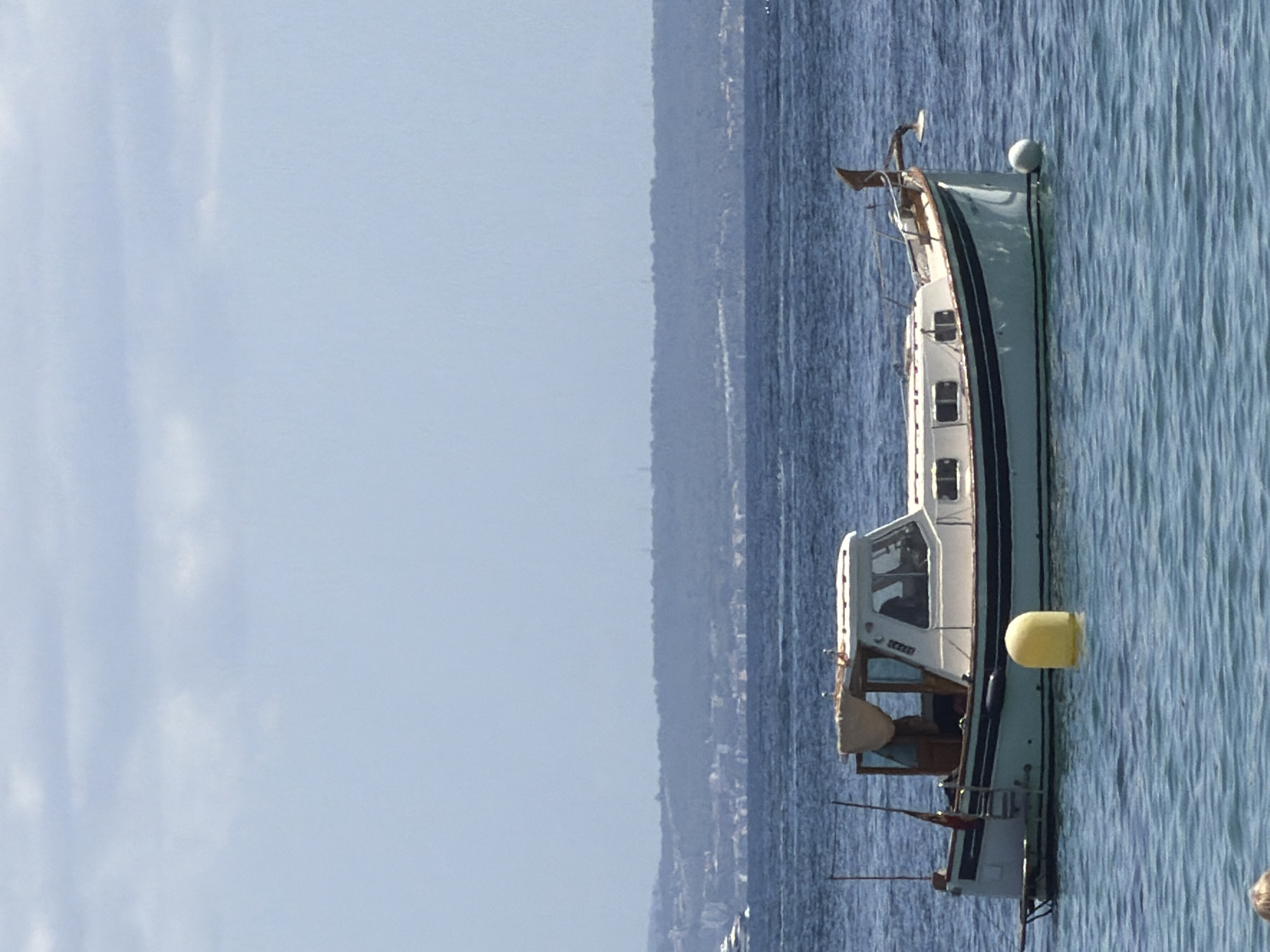 A motor yacht, approximately 8-10 meters in length, is anchored in the blue water off the coast of Palmanova, Spain. The yacht is white with a dark blue stripe along its waterline and a beige canopy over the main cabin. It has several windows, a ladder leading up to the deck, and a flag at the stern. A yellow buoy floats near the yacht's stern, and a white fender is attached to its port side. In the background, a hazy, green-covered hill with distant buildings and some structures on top is visible. The sky is light blue with scattered white clouds. The water is slightly choppy. There are no visible people on the yacht or in the immediate vicinity in the water. The scene appears peaceful and calm.