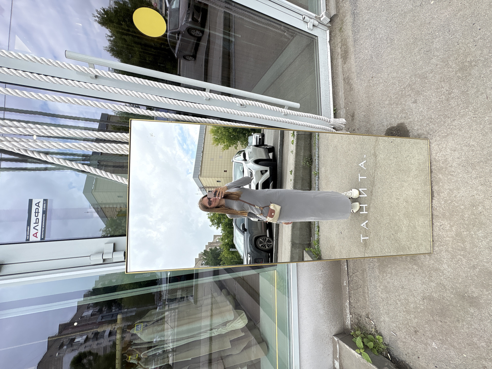 A young woman is taking a selfie in a large, full-length mirror positioned outdoors in front of a building with a glass facade. The mirror reflects the woman, who is wearing a long, light gray dress, white sneakers, and sunglasses. She is holding a pink smartphone in her right hand. A small, two-toned handbag is slung across her body. The mirror is framed in gold and has white rope decorations hanging from the top. In the reflection, the woman is standing on a concrete sidewalk. Behind her, cars are parked along a street. The building's glass facade reflects the sky, trees, and other buildings. A sign on the glass facade reads "НА С ОХРАНЯЕТ АЛЬФА", which translates to "IS PROTECTED BY ALPHA". The surroundings suggest an urban environment, possibly a commercial street. Based on the text on the sign, the location is in the city of Perm, Russia.