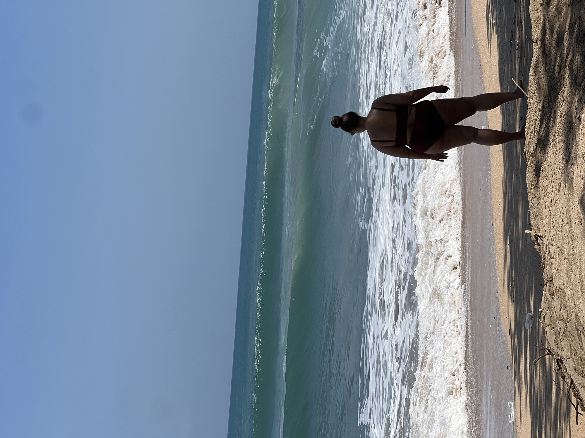 The image shows a person wearing a dark red bikini standing on a sandy beach, facing the ocean. The person has their hair styled in a bun. The ocean has rolling waves and is a turquoise color. The sky is clear and blue. There is some litter on the sand in the foreground, including plastic bottles. The location is the city of Kuta, Indonesia.