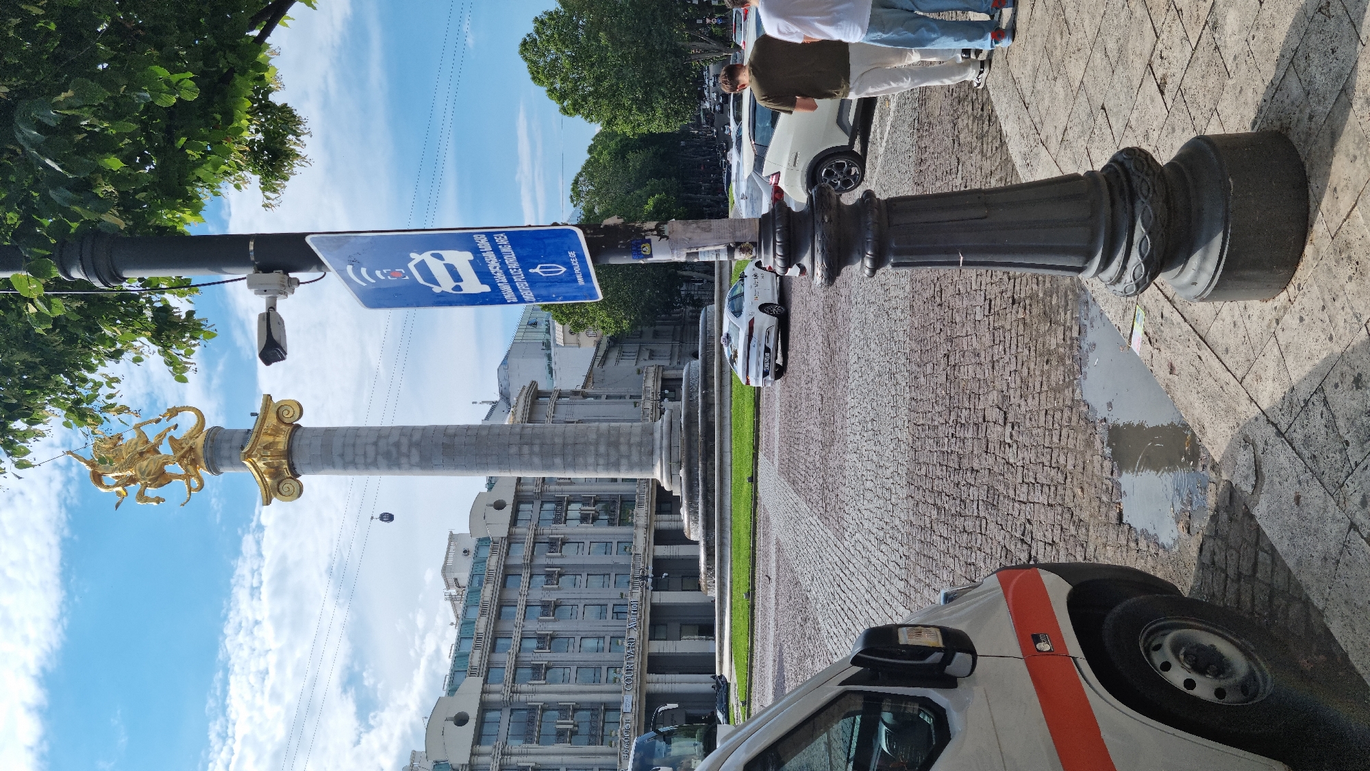The image shows a street scene in Tbilisi, Georgia, likely in the vicinity of Freedom Square, given the prominent monument. In the foreground, a white van with an orange stripe is parked on the left side of the frame. A black ornate lamppost stands to the right, with a blue sign attached to it indicating a "PATROLLED POLICE AREA" with a police car icon and a QR code. A security camera is mounted on the lamppost above the sign.
In the mid-ground, a white car, possibly a taxi, is driving on the cobblestone street. Behind it, a large circular monument with a golden statue of Saint George on horseback adorns the top, supported by a tall stone column. The monument is situated in a large open plaza with a fountain at its base, surrounded by a green lawn. In the background, a grand building with classical architecture, identified by signage as the "Courtyard Marriott," is visible. Several trees line the street, and a cable car is suspended in the sky.
Two individuals are partially visible in the lower right corner of the image, standing on the sidewalk. The one on the left is wearing a dark green t-shirt and light-colored trousers, while the one on the right is wearing a white t-shirt and blue jeans. They appear to be looking at their phones. The sky is partly cloudy. The street is paved with cobblestones, and a puddle of water is visible on the ground near the van and lamppost.