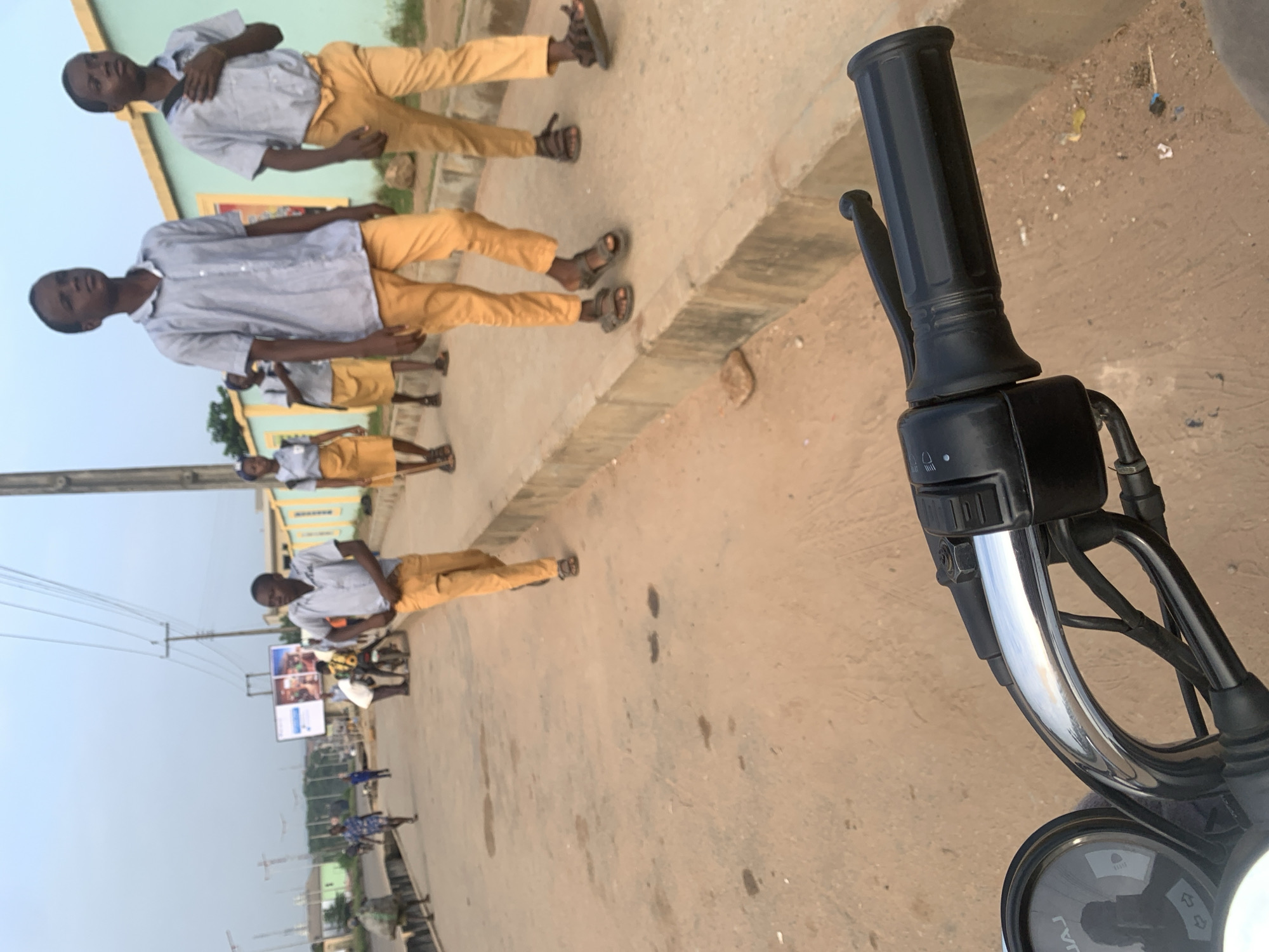 The image depicts a street scene in Shagamu, Nigeria, viewed from the perspective of someone on a motorcycle. In the foreground, the handlebars and part of the dashboard of the motorcycle are visible. The road is unpaved and dusty, with a concrete curb running along the right side. A group of young people, likely students in uniform, are walking on the sidewalk and the edge of the road. They are wearing light blue shirts and yellow trousers. Some are walking in pairs or small groups. In the background, there are buildings, utility poles with electrical wires, and a billboard. Other individuals are also present on the street, including someone carrying a large bag and a person on a motorcycle. The sky is overcast, suggesting it is either early morning or late afternoon. The overall impression is one of a typical urban environment in Nigeria.