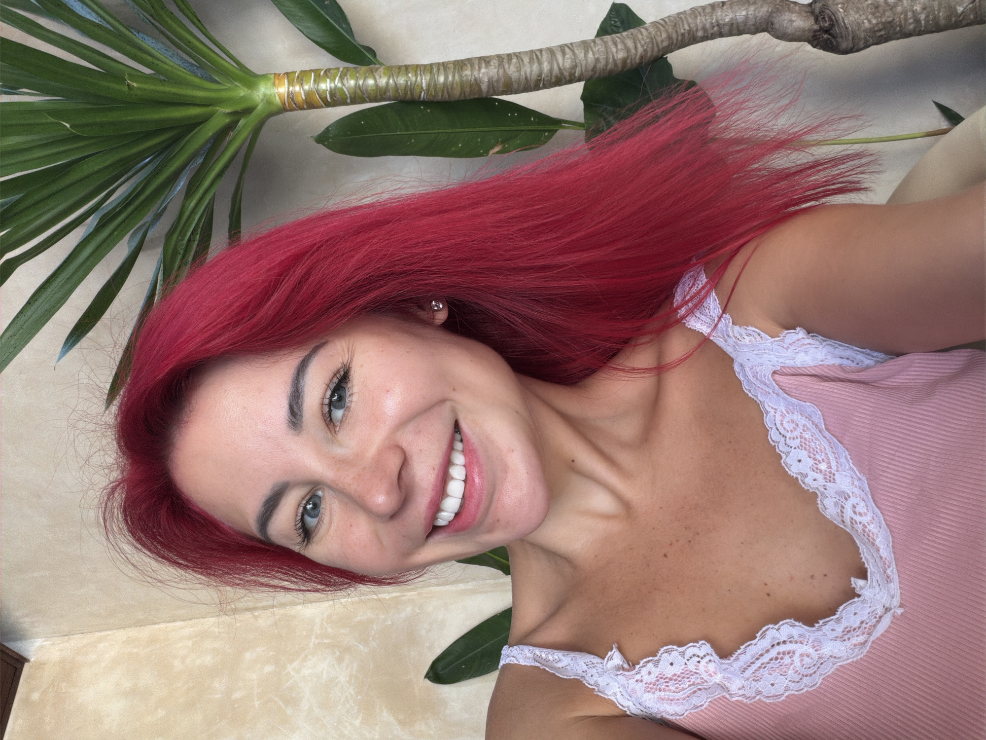 A woman with bright red hair is captured in a selfie. She is wearing a pink camisole with white lace trim. She has blue eyes and a wide smile, showing her teeth. Freckles are visible on her cheeks and chest. Behind her, a plant with large green leaves and a textured trunk is present. The background appears to be a wall with a textured, light-colored finish. The setting is indoors. The question about Kuta, Indonesia is irrelevant to the visual content of the image.