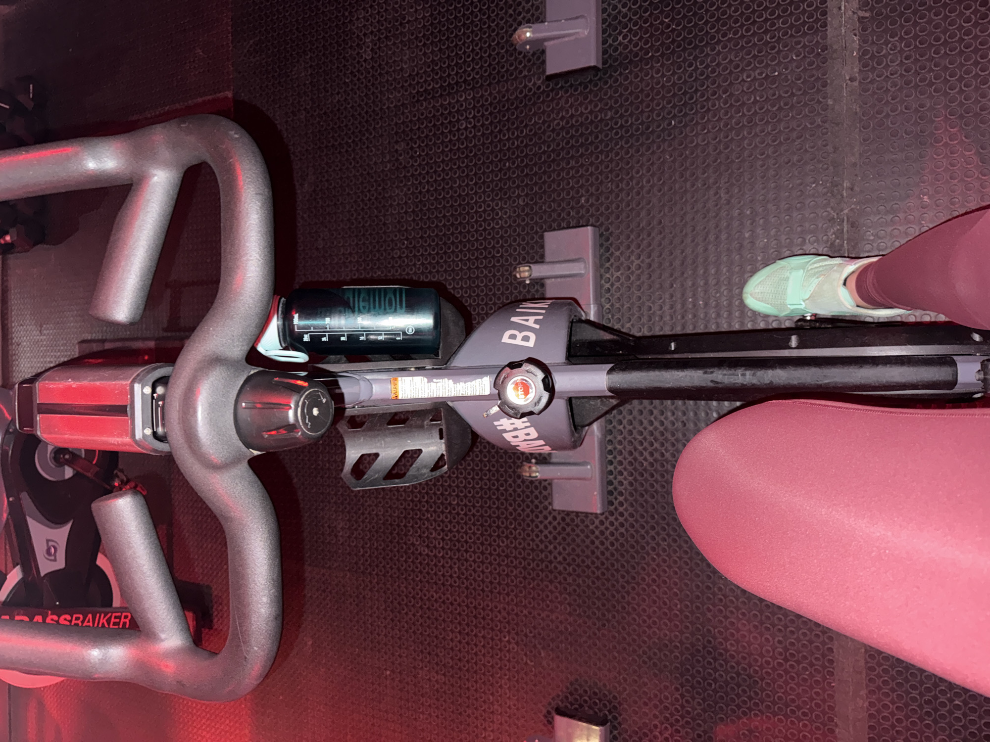 The image shows an indoor cycling bike, viewed from above. The handlebars are silver and curved, with a digital display screen. The bike's frame is dark gray with a red accent and the word "CROSSRAIKER" visible. Pedals are visible. A person is seated on the bike, wearing burgundy leggings and a light blue sneaker. The environment is a gym with a textured black floor. There is no information in the image or requested that mentions Puebla, Mexico.