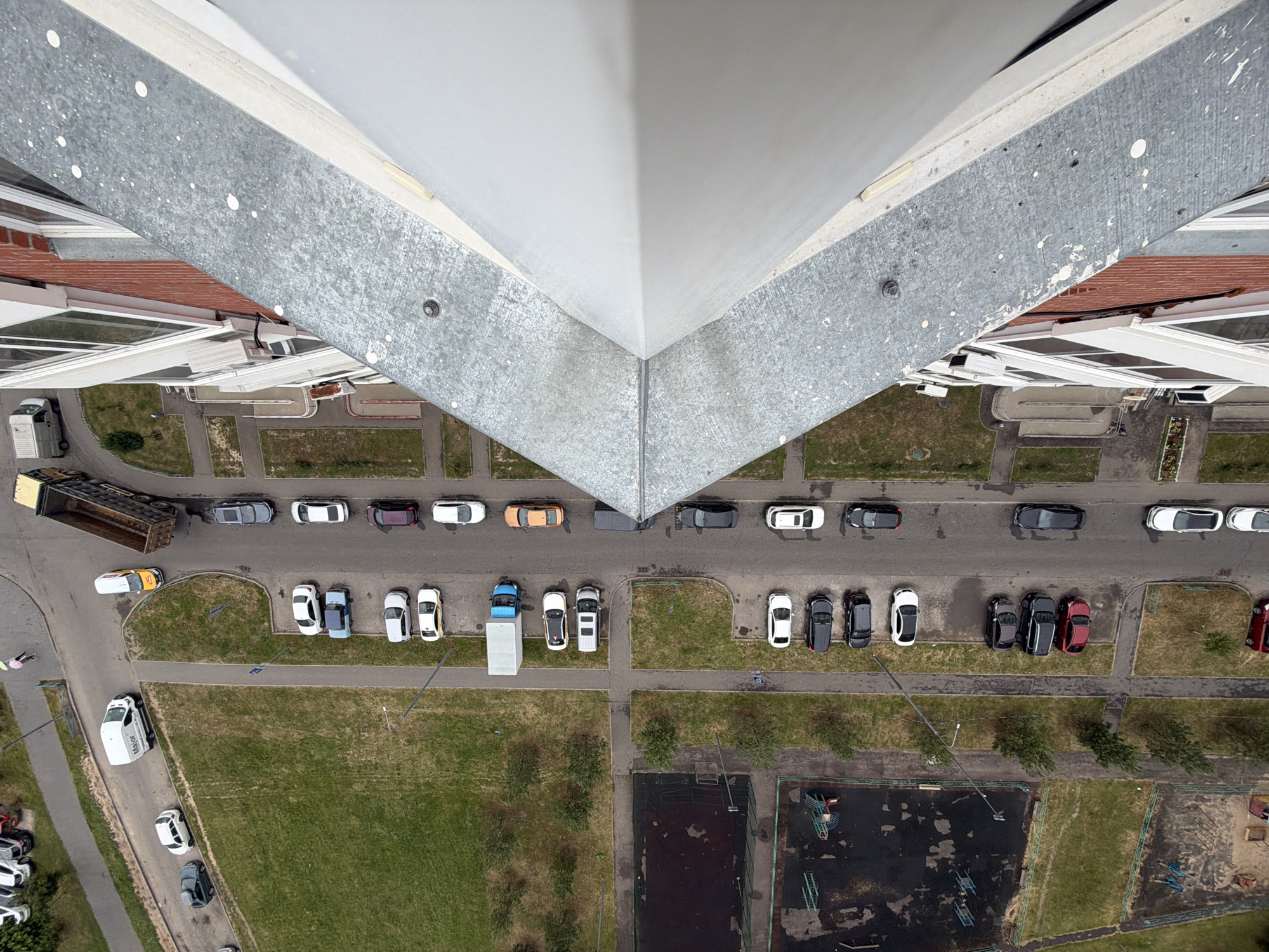 This is an aerial, high-angle shot of a residential area in Bobrovo, Russia. The perspective is looking down from a significant height, with the sharp, angular roofline of a building dominating the top portion of the frame. Below the building, a street and parking area are visible, lined with parked cars of various colors and models. A large, empty dump truck is parked on the side of the road, its bed tilted. Some green spaces with grass and scattered trees are interspersed between parking areas and the buildings.
In the lower right quadrant of the image, there appears to be a sports court or playground area, with dark, worn surfaces and some playground equipment visible. A few people are faintly visible in the scene, one walking on the road and another near the edge of the grassy area. The time of day appears to be daytime, with adequate lighting for visibility, suggesting it is not early morning or late evening. The weather seems clear, with no indications of rain or overcast skies. The scene depicts a typical urban or suburban environment, likely on a weekend or a non-working day given the number of parked cars and the relatively quiet scene. No discernible text is visible on any of the objects or structures in the image.
