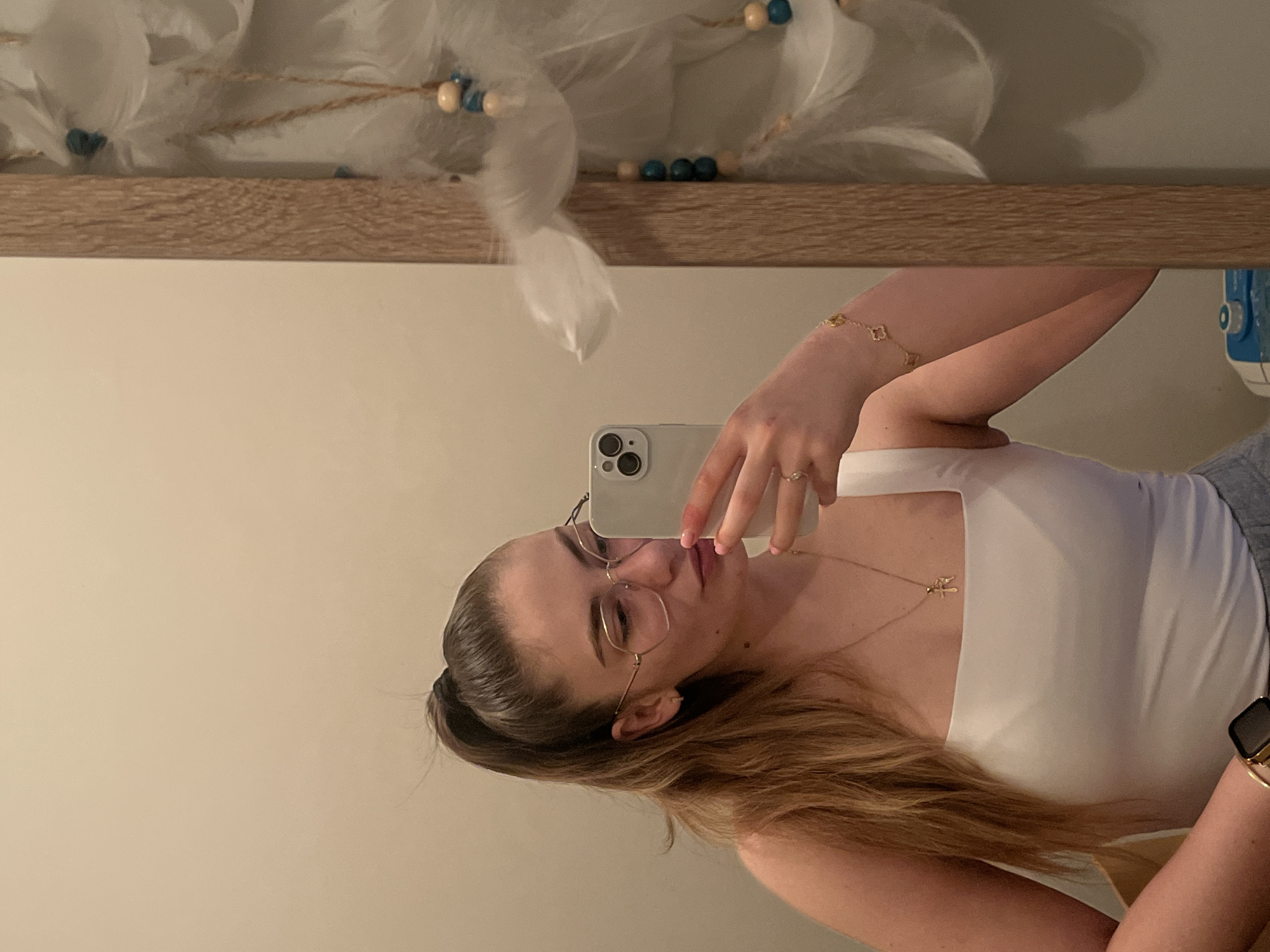 A young woman is captured in a mirror selfie, her image rotated 90 degrees counter-clockwise in the frame. She has long, light brown hair styled in a high bun or ponytail and wears thin, gold-rimmed glasses. Her expression is neutral with a slight hint of a smile.
She is dressed in a white tank top and adorned with several pieces of jewelry: a delicate gold-toned necklace with a small pendant, possibly a cross; a gold-toned bracelet featuring small clover-shaped charms on her left wrist; and a smartwatch on her right wrist. Rings are visible on her left ring finger and right index finger, and her fingernails are painted a light pink or nude color.
The woman is holding a light-colored smartphone, possibly an iPhone given the camera module design, which reflects her face. The selfie was taken indoors, with a plain, light-colored wall serving as the background in the mirror's reflection. Above this wall, a light-colored wooden shelf or frame runs horizontally. Resting on or hanging above this shelf are decorative elements resembling white feathers, woven with light brown string or rope, and interspersed with small, round beads in shades of blue and off-white. There is no visible text in the image.