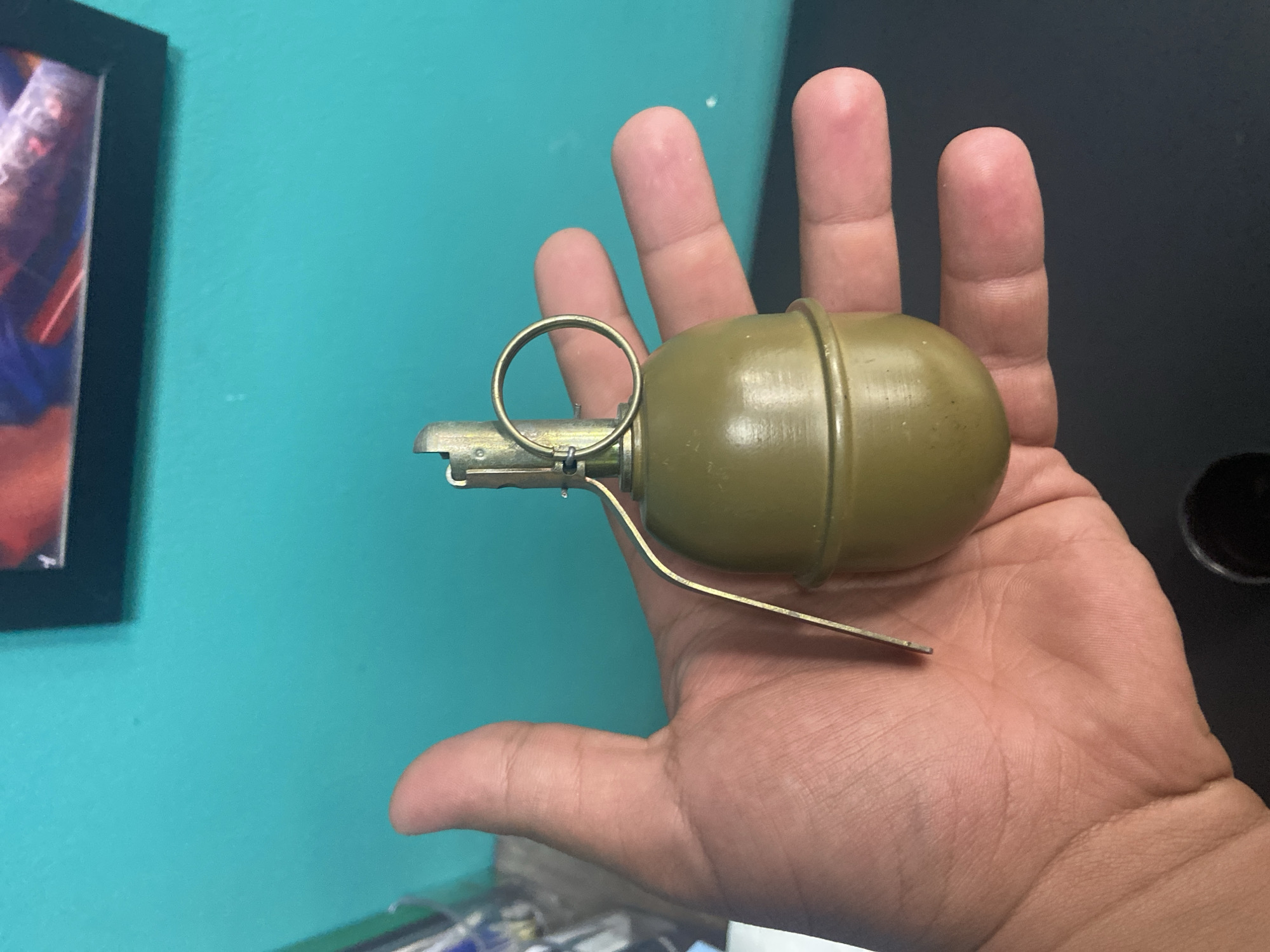 A person's hand is holding a green, oval-shaped object with a metal lever and ring, resembling a hand grenade. The background consists of a turquoise wall with a framed picture at the top and a dark surface below. The image appears to be taken indoors. There is no information that can be used to determine the location in the city of Yegorlykskaya, Russia.