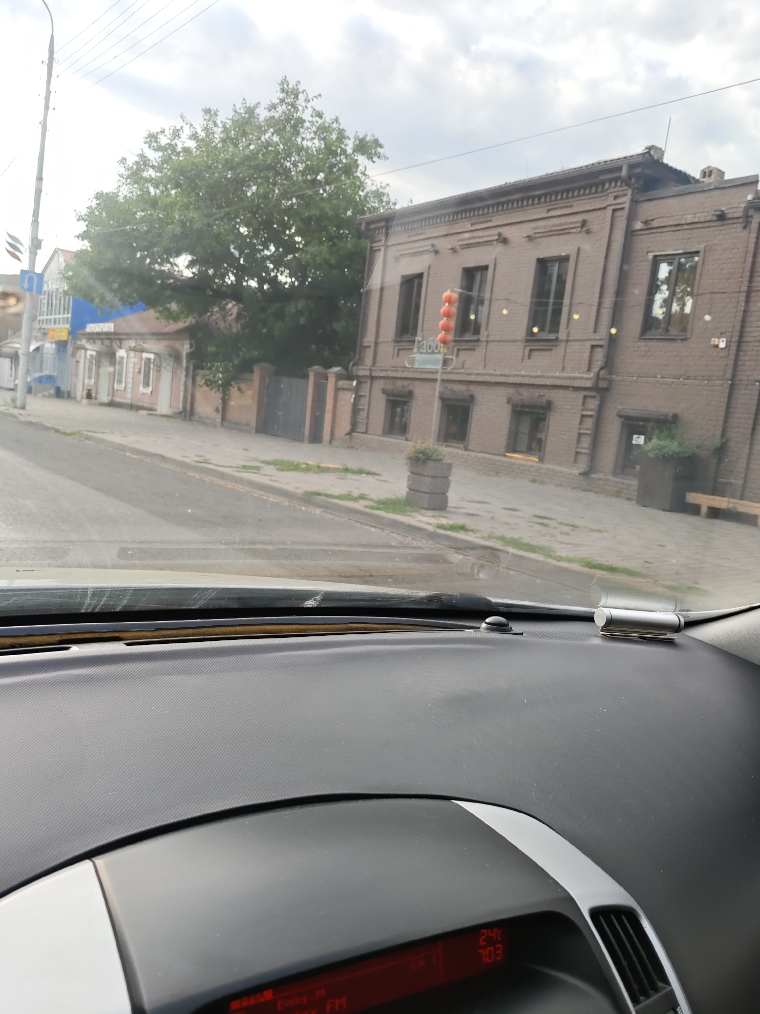 The image shows a view from inside a car, looking out at a street scene in Novocherkassk, Russia. The car's dashboard is in the foreground, with a digital display showing "24°C" and "7:03". Outside, a brick building with multiple windows and an arched entrance is visible, adorned with hanging lights. Across the street, there are other buildings, including a blue one with windows, and a large tree. The street is paved, with sidewalks and grassy areas along the side. A traffic light is visible on a pole. The sky is overcast.