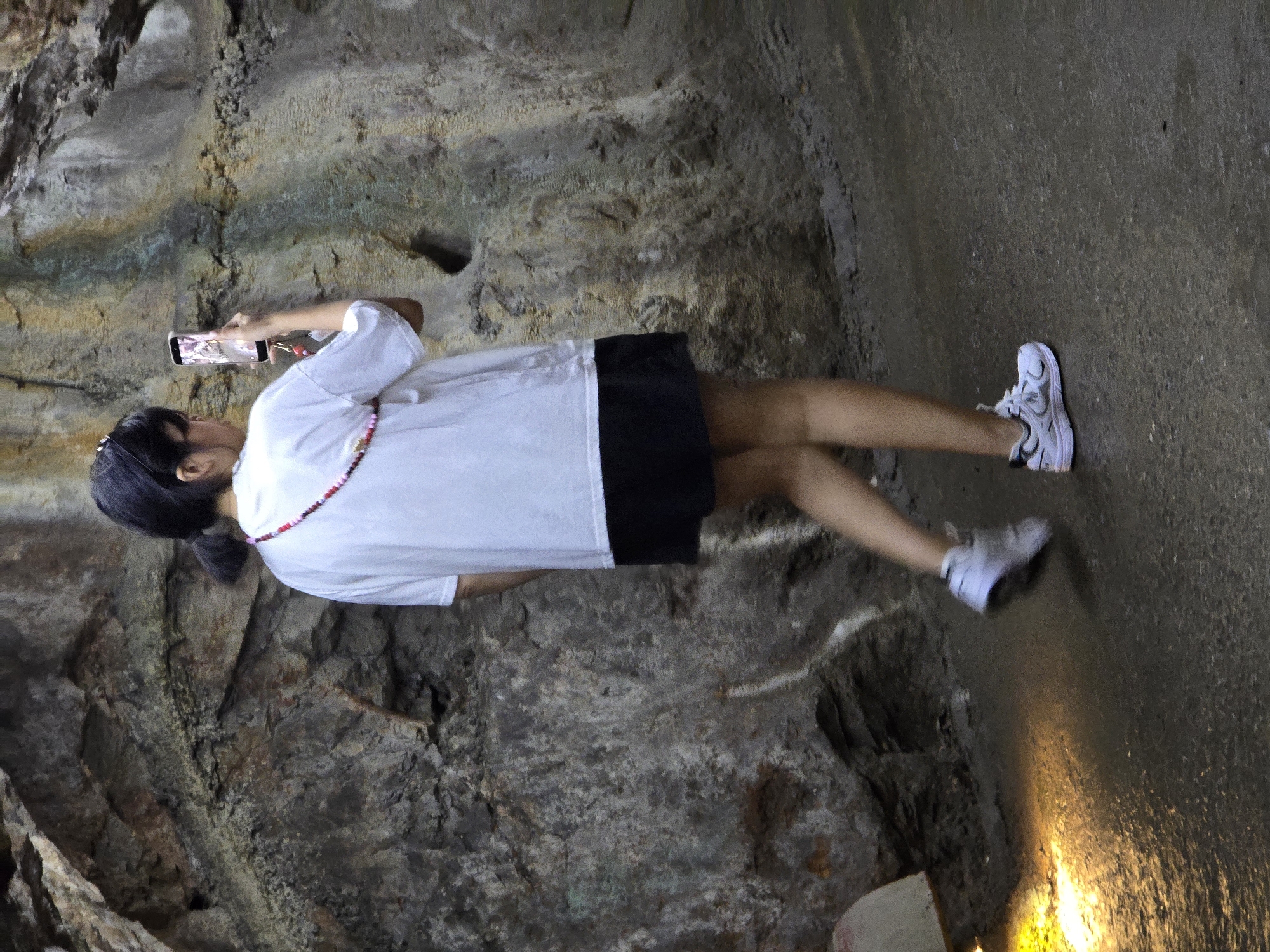 A young Asian female wearing a white t-shirt and black skirt is captured from behind as she takes a photograph with her cell phone. The woman's hair is pulled back into a ponytail, and she has a colorful beaded lanyard around her neck. She is standing on a wet, dark-colored path in what appears to be a cave or a rocky, natural environment. The wall behind her is made of rough, textured rock with streaks of yellow and green. A bright yellow light shines on the wet ground to her left. Her white sneakers are visible as she stands with her body angled slightly away from the viewer.