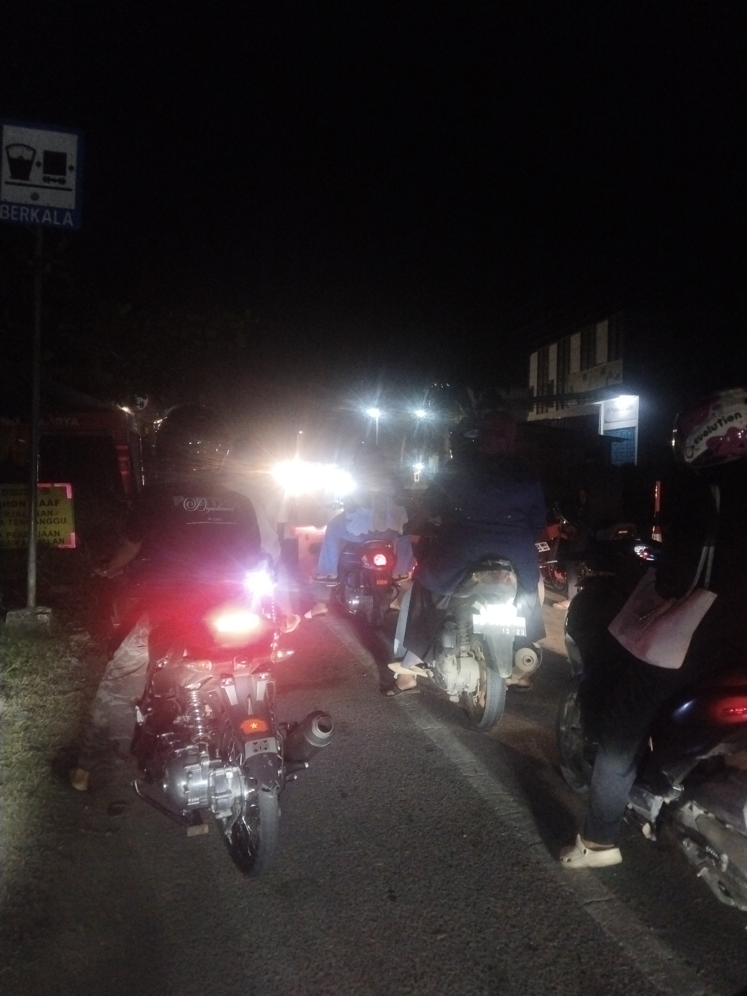 The image shows a congested street scene at night in Lhokseumawe, Indonesia. Several motorcycles are lined up on the road, facing forward. People are seated on some of the motorcycles, likely waiting for traffic to move. A sign post with a white rectangle and the word "BERKALA" in a blue field is on the left. The headlights of an unseen vehicle illuminate the scene. A sign partially readable with the word "Departemen" is on the left side. The number "12-23" is visible on one of the motorcycles. A building with lit windows is in the background.