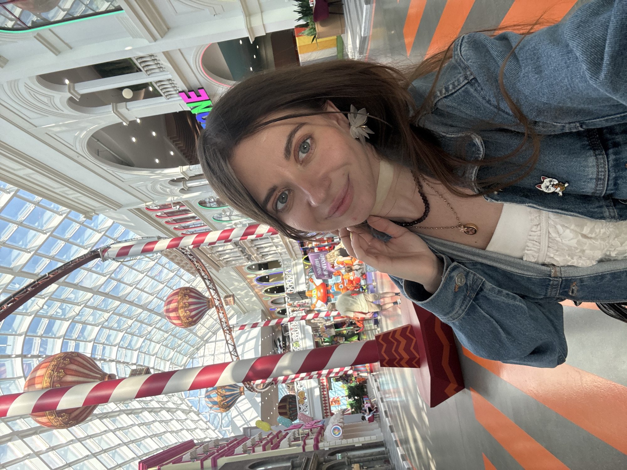 A medium-angle shot depicts a woman taking a selfie in an indoor shopping mall environment, likely in Moscow, Russia, given the architectural style. The woman, with long brown hair and wearing a denim jacket, faces the camera with a slight smile. She has light skin and wears a small, decorative flower-like object near her ear. She is wearing jewelry including necklaces and a pin.
The background reveals an elaborate shopping mall interior with a high, grid-patterned glass ceiling, suspended decorative hot air balloons, and candy cane-striped architectural elements. In the distance, there are stores and displays, and a few people can be seen shopping. The floor has a distinct orange and gray striped pattern. The lighting is bright, suggesting natural daylight filtering through the glass ceiling.