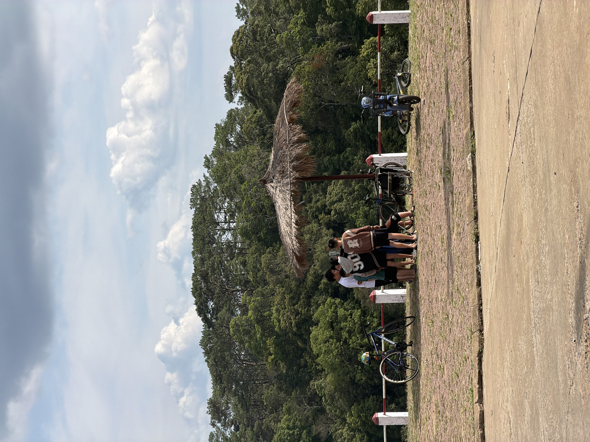 The image depicts an outdoor scene, likely a rest stop, under a bright sky with scattered white clouds, situated in a densely forested, mountainous region. The foreground features a wide, light-colored concrete road on the right, separated by a distinct crack from a brick-paved path on the left.
In the midground, a group of approximately five to seven young people, mostly males, are gathered under a large, round thatched umbrella, providing shade on the brick pathway. They are dressed in casual attire, including t-shirts and shorts, and are largely seen from behind, suggesting they are conversing or resting. One individual wears a shirt with the number "06" visible on the back.
Several bicycles are parked nearby. A prominent blue bicycle, with a helmet resting on its seat, leans against a white and red striped barrier post on the left side of the brick path. Another dark-colored bicycle is visible further along the path to the right of the group, also near barrier posts. These barriers delineate the edge of the brick pathway.
The background is dominated by a lush, green forest composed of numerous coniferous trees, extending across the horizon under the bright sky. The strong sunlight and clear skies indicate it is daytime, likely mid-morning or early afternoon. The scene suggests a leisure activity, possibly a cycling trip, where the participants have paused for a break in a scenic location, consistent with the context of Măng Đen, Vietnam.