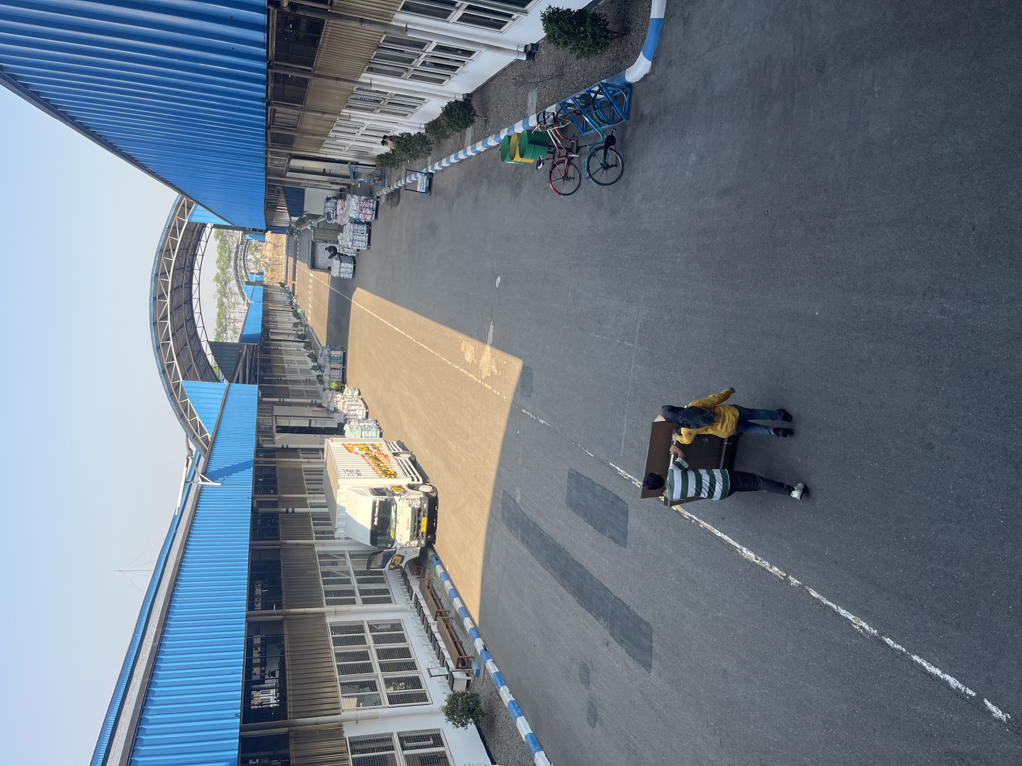 The image depicts an outdoor scene at a logistics or distribution center in Pelang, Indonesia. The architecture features long, blue-roofed buildings with numerous windows, likely warehouses or factory units, connected by covered walkways. Sunlight casts long shadows across the asphalt pavement, indicating it is likely daytime.
In the foreground, two individuals, a man and a woman, are pushing a large cardboard box. The man is wearing a striped green and white t-shirt and dark pants, while the woman is wearing a yellow long-sleeved shirt and blue jeans. They are walking away from the camera, towards the right side of the frame.
Further into the scene, a white truck with some branding visible is parked. Several pallets stacked with goods, possibly merchandise or supplies, are lined up along the buildings. In the middle ground, there are two bicycles parked near a blue and white striped curb, alongside some trash receptacles. In the background, the covered walkways continue, leading to other structures and what appears to be vegetation in the distance. The overall impression is one of industrial activity and goods movement.