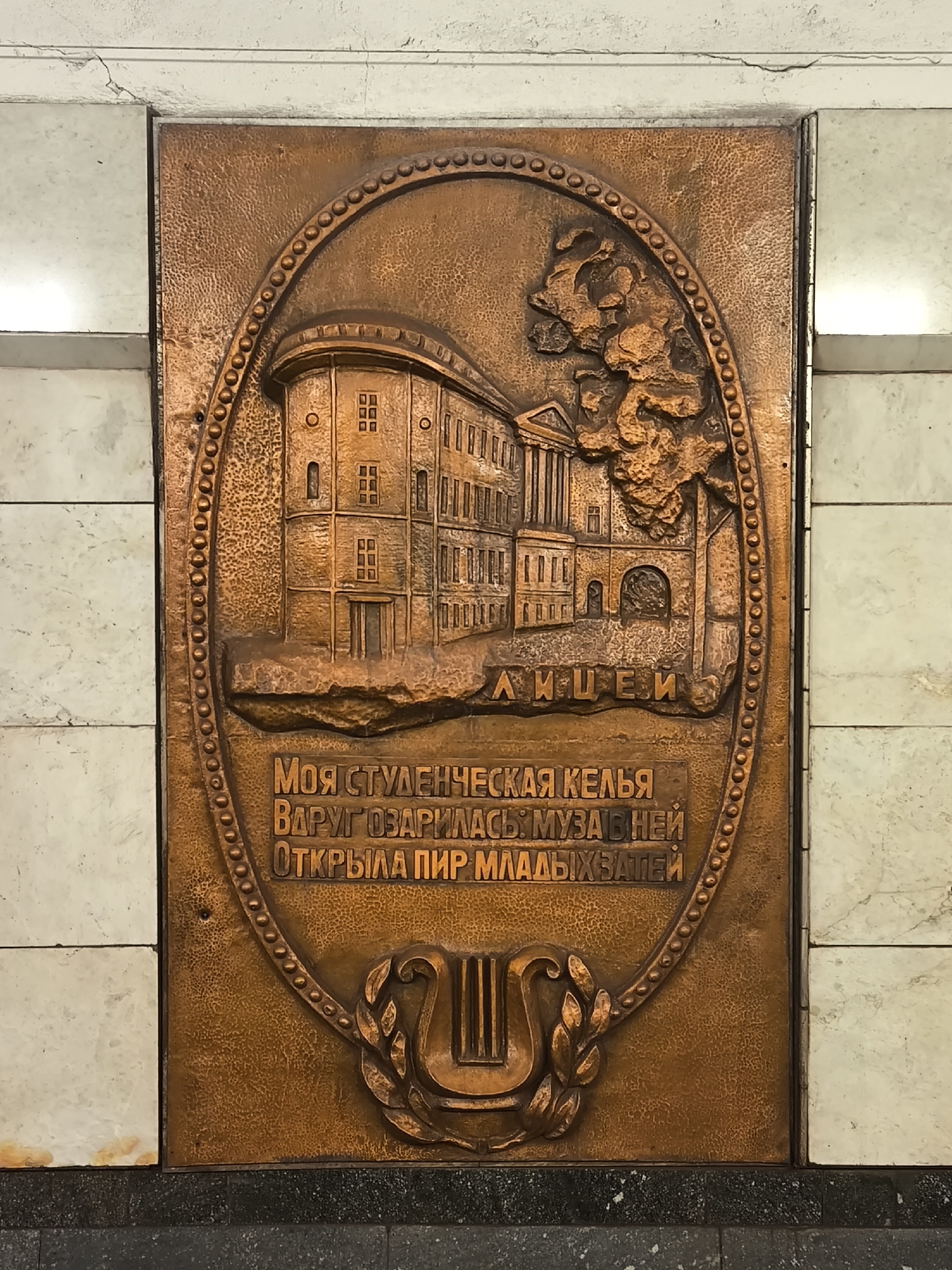 This is a close-up of a copper relief panel located at the Komsomolskaya-Radialnaya metro station in Moscow, Russia. The panel is rectangular with an oval centerpiece. The centerpiece contains a bas-relief depicting the facade of a classical building with the word "ЛИЦЕИ" (Lyceum) beneath it. Below the building is an inscription in Russian: "МОЯ СТУДЕНЧЕСКАЯ КЕЛЬЯ ВДРУГ ОЗАРИЛАСЬ: МУЗА В НЕЙ ОТКРЫЛА ПИР МЛАДЫХ ЗАТЕЙ." At the bottom of the oval is a lyre with laurel branches. The panel is mounted on a wall with white marble tiles above and dark gray tiles below.