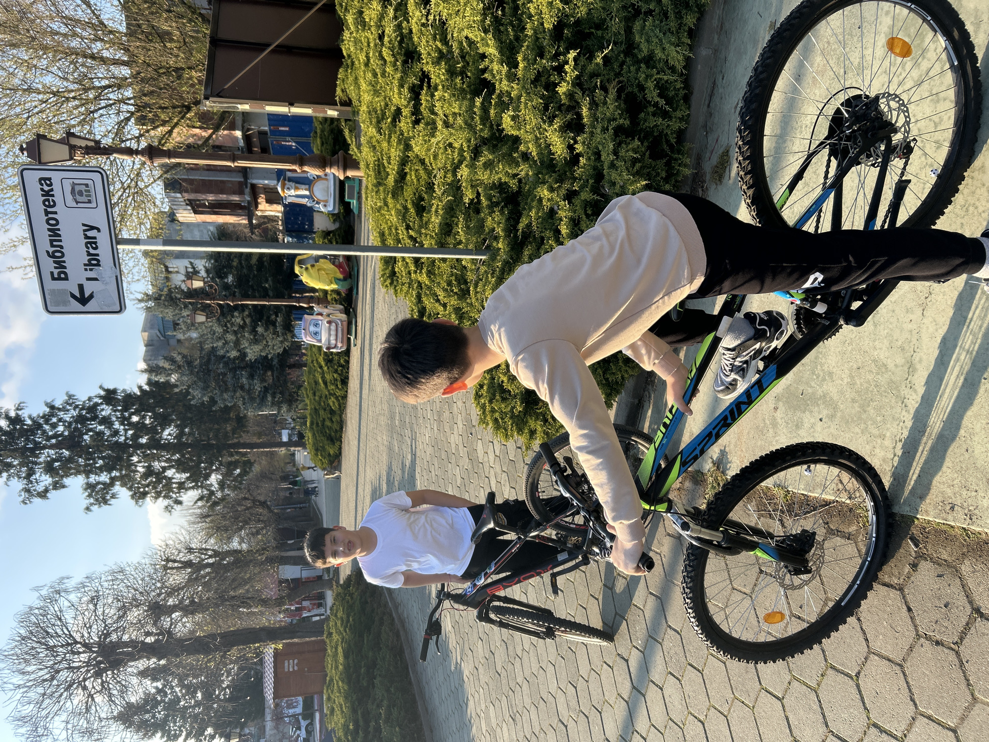 Two young males are standing in a paved outdoor area next to bicycles. The male closer to the camera is wearing a white T-shirt and dark pants, holding his bike by the handlebars. The second male is wearing a light-colored long-sleeve shirt and dark pants. He is leaning over his bicycle, appearing to adjust something. The bicycles are mountain-bike style, with visible branding on one that reads "SPRINT".
A signpost indicating "Biblioteca / Library" in both Bulgarian and English is visible in the background. There are also decorative elements such as street lamps and small ride-on toys for children. Evergreen bushes and bare trees are present in the background. The location is Zlatograd, Bulgaria.