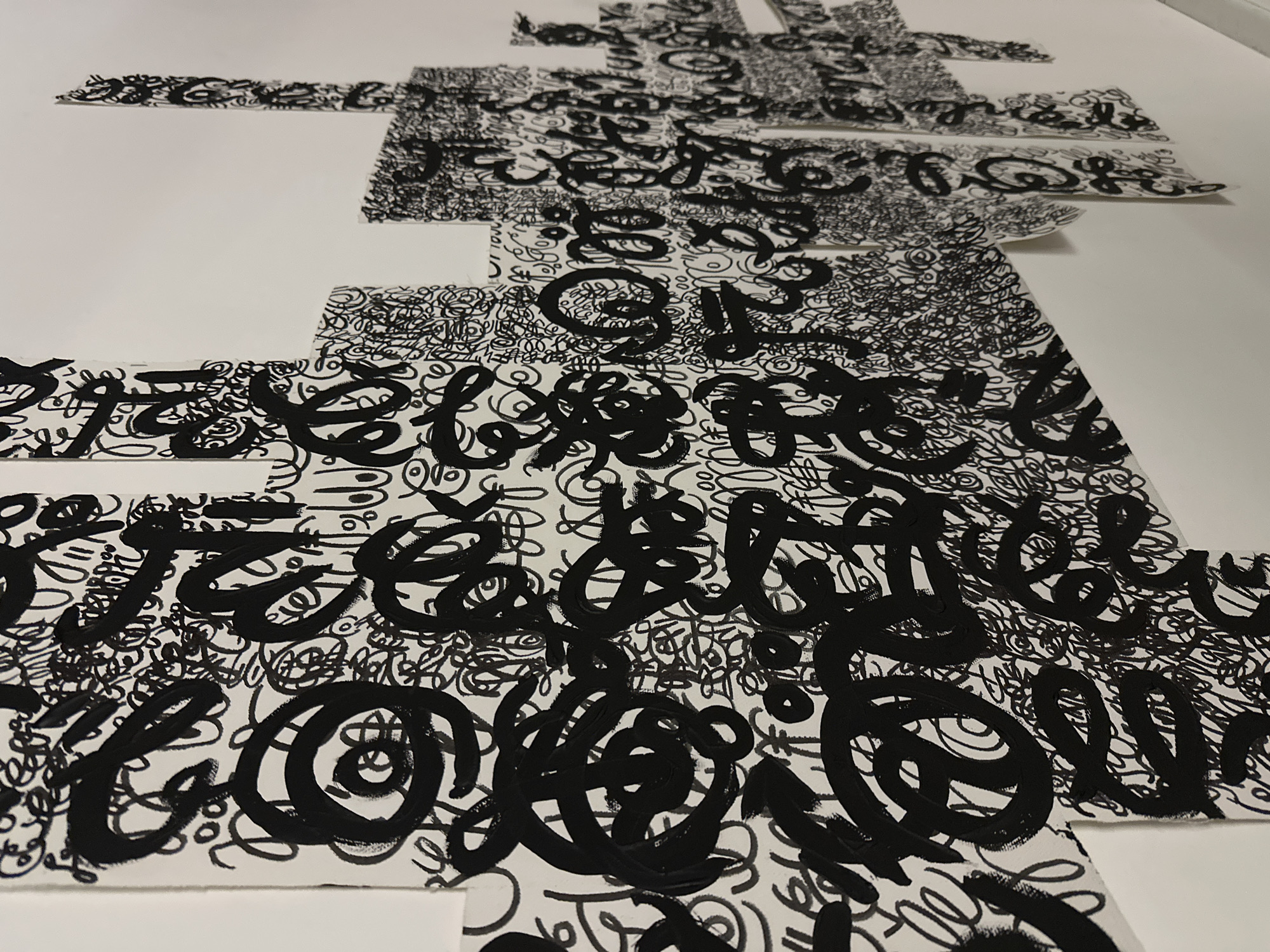 A close-up, high-angle shot displays segments of a large, abstract art piece, potentially a collage or assemblage, made of paper or similar material. The artwork features dense, black, scribbled lines and calligraphic elements against a white background. The black lines vary in thickness, some appearing bold and painted, while others are finer, like ink. The segments of the art are arranged with slight overlaps and spaces between them, creating a fragmented, layered effect. The overall composition has a dynamic, chaotic energy due to the density and randomness of the markings. The scene is situated in a well-lit environment, possibly a studio or gallery space. The location in Moscow, Russia, is not discernible from the image alone. There are no people visible, and the environment is primarily the immediate surroundings of the artwork.