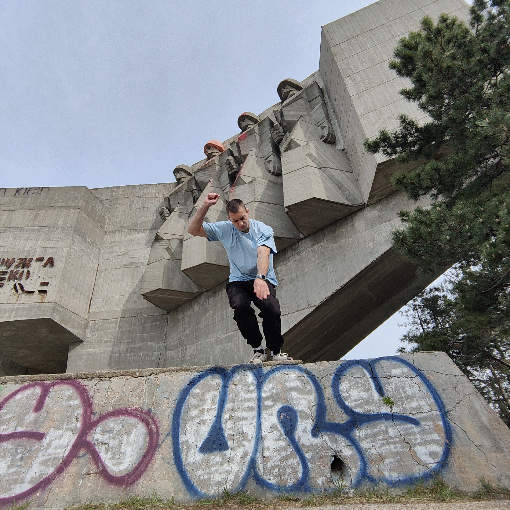 A low-angle shot depicts a man jumping on a graffiti-covered concrete structure in front of a large brutalist-style monument in Varna, Bulgaria. The monument is made of concrete and features stylized figures with headgear. The man is wearing a light blue t-shirt and dark pants. Graffiti is visible on the lower part of the concrete structure. A tree with green foliage is present on the right side of the frame. The sky is overcast.