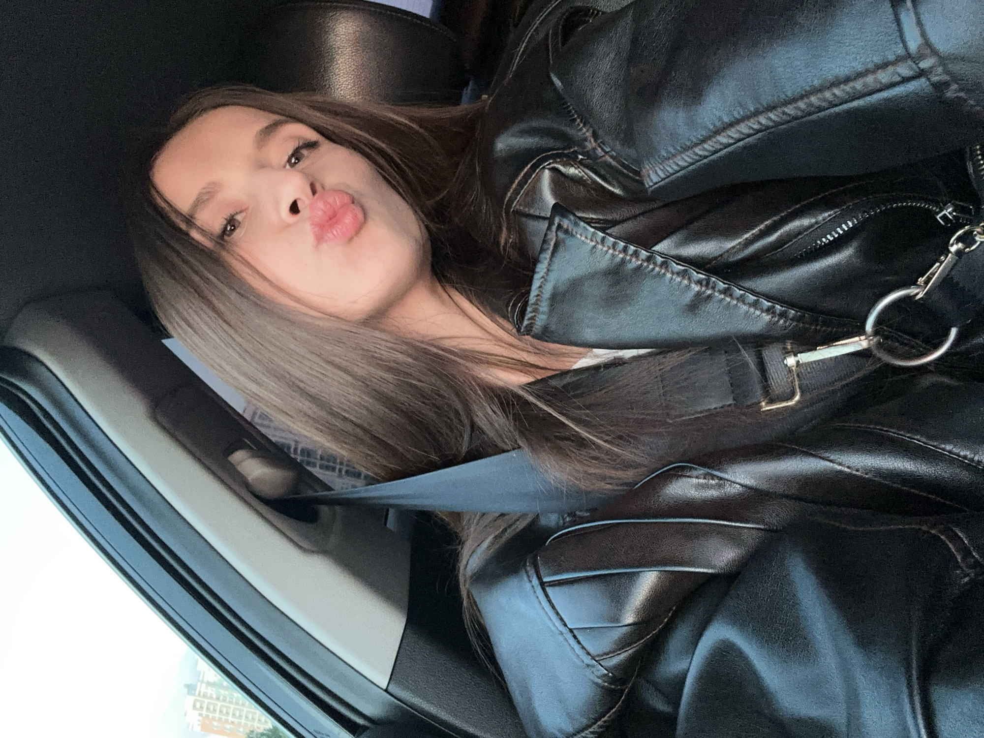 A young woman is captured in a close-up selfie inside what appears to be a car, possibly in Yekaterinburg, Russia. She has long, straight brown hair that cascades over her shoulders and part of her chest. Her eyes are open, looking towards the camera, and she has subtle eye makeup. Her lips are puckered into a "kissy face" expression, indicating a playful or affectionate mood.
She is wearing a shiny black leather jacket, which is prominent in the frame, showcasing details like zippers and stitching. A black seatbelt is visible across her chest and shoulder, indicating she is seated in a moving or parked vehicle. The interior of the car is dark, possibly with black leather seats and trim.
Through the car window on the left, a blurry background suggests an urban environment with buildings, consistent with a city setting like Yekaterinburg. The lighting indicates it is daytime, although the specific weather conditions are not clearly discernible from the interior perspective. The overall scene conveys a casual moment captured during travel, with the subject engaging directly with the camera. No visible text is present in the image.
