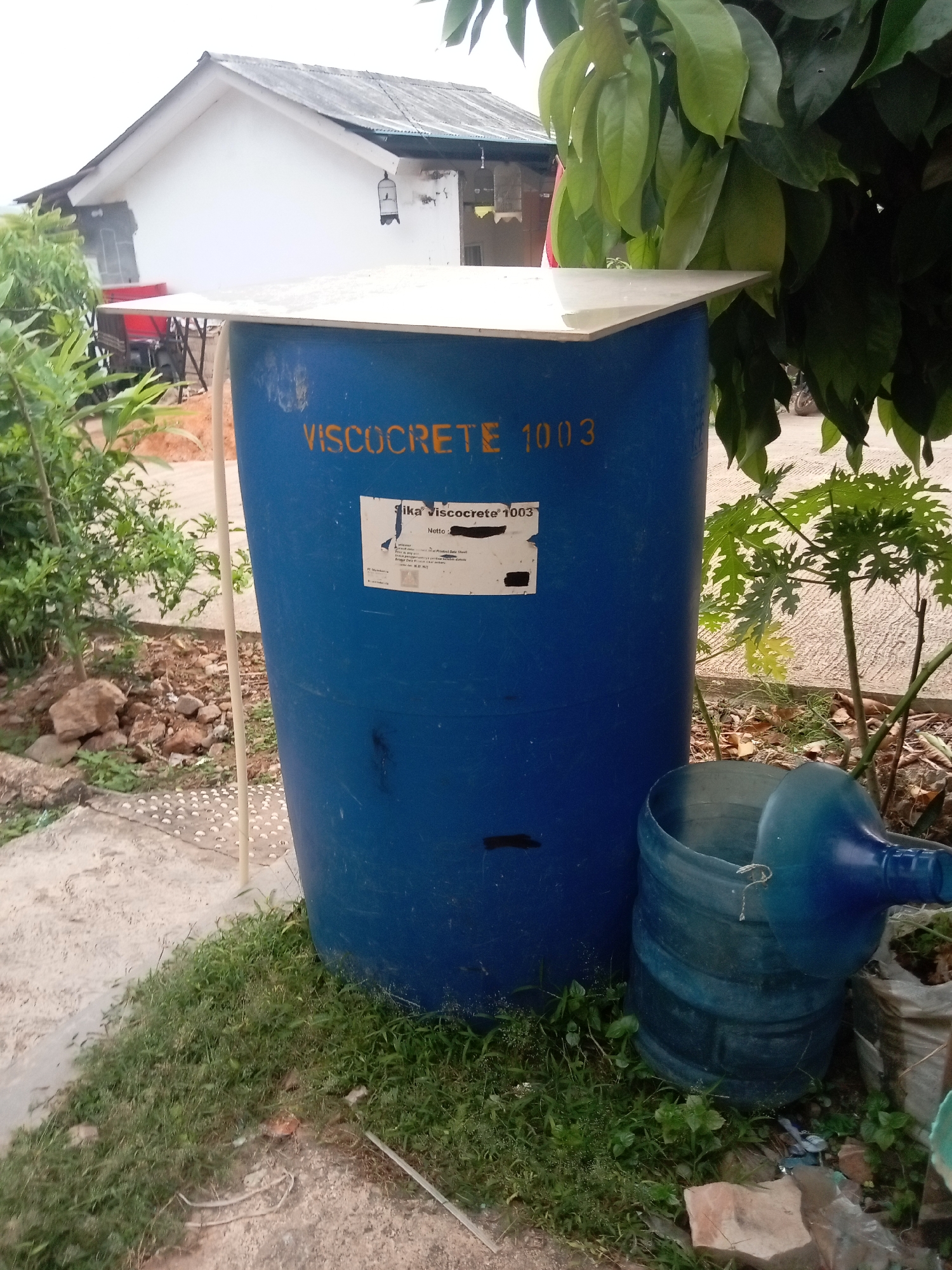 The image depicts an outdoor scene near a residential building in Batam, Indonesia. The primary subject is a large, blue plastic drum with "VISCOCRETE 1003" printed in yellow on its side. A white label with text is affixed to the drum. To the right of the drum is a large, blue plastic water jug with its top removed, revealing the interior. The drum is situated on a patch of grass, and behind it, a part of a white house with a gray roof is visible. There are green plants and trees surrounding the drum and jug. The ground appears to be a mix of dirt and gravel. A white pole supports the lid of the blue drum. The overall setting suggests a storage or utility area in a residential neighborhood.