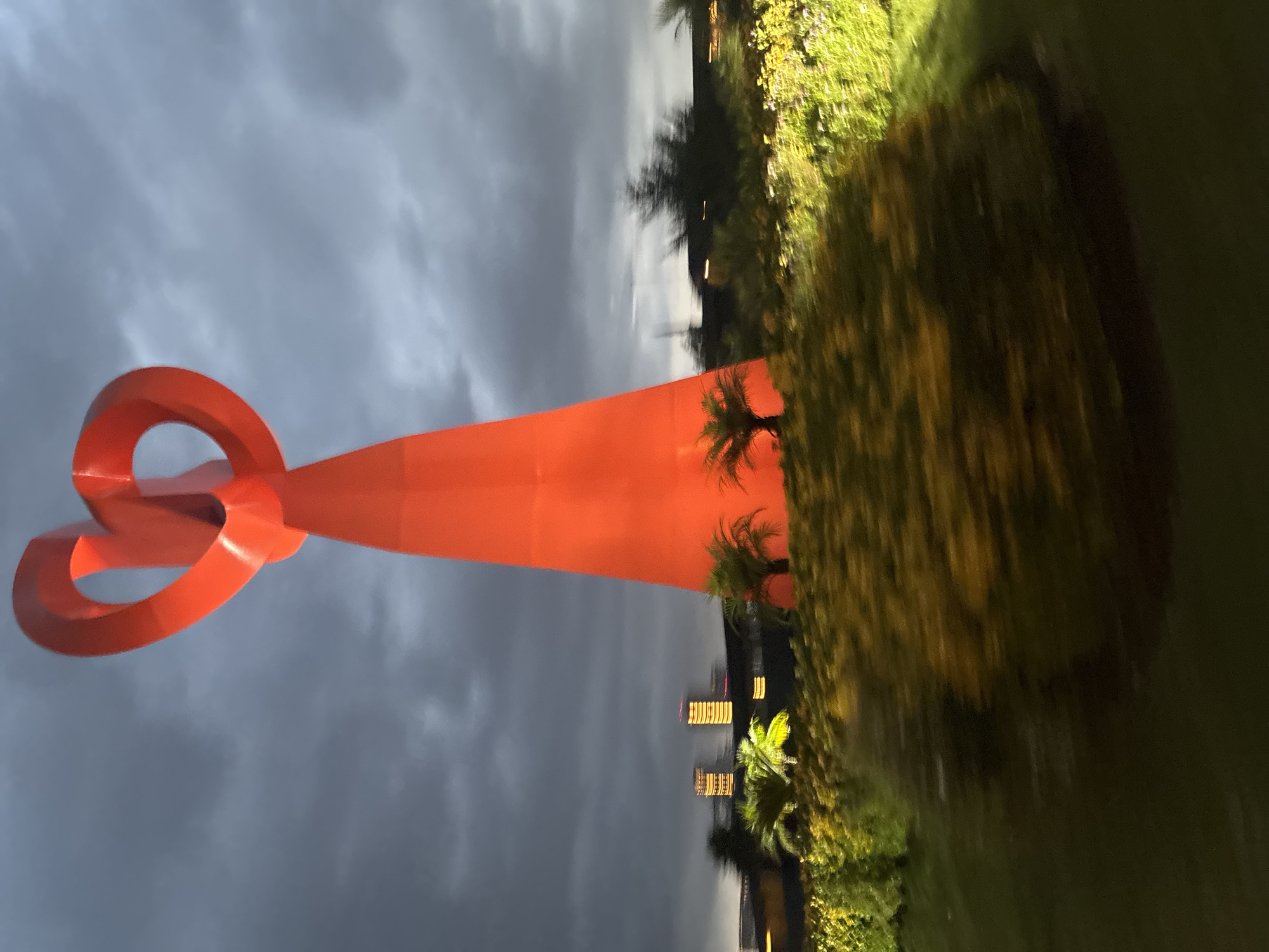 The image shows an outdoor scene featuring a large, orange, geometric sculpture. The sculpture consists of a long, tapered rectangular form that culminates in a trio of interconnected circular loops. The sculpture is located in Huixquilucan de Degollado, Mexico. The background features a cloudy sky transitioning to a cityscape with lighted buildings and green vegetation, including palm trees. The foreground consists of shrubs and trees along the ground. The lighting is dim, suggesting it is dusk or evening. The photograph is rotated ninety degrees counterclockwise.