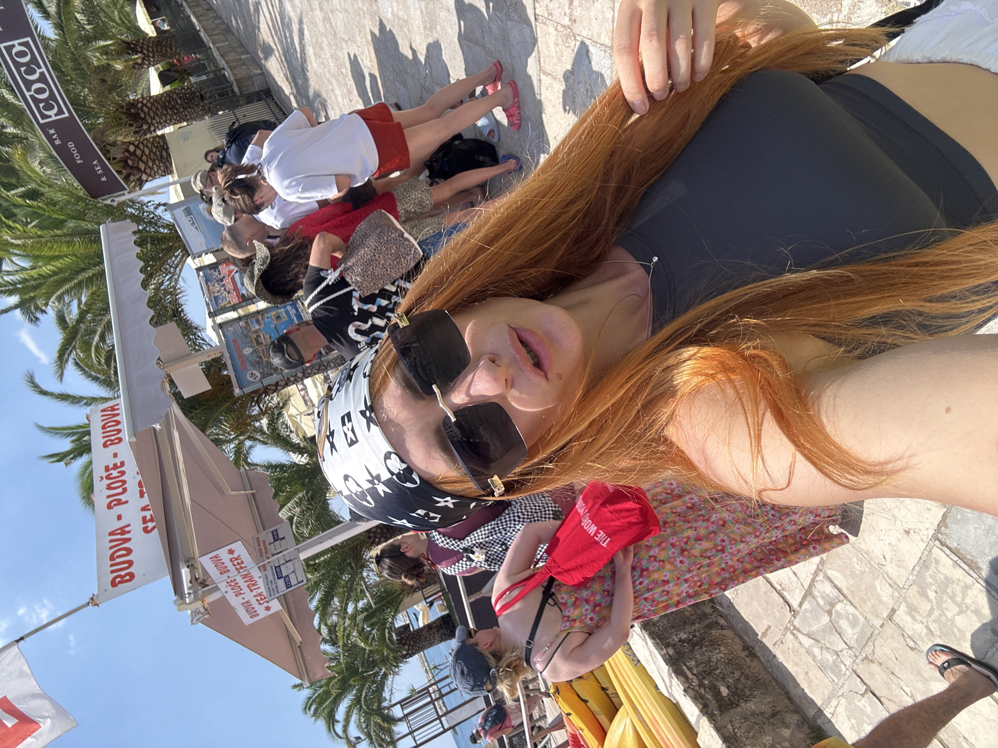 This media file captures a lively outdoor scene in Budva, Montenegro. The focus is on a group of people, with a woman with long, reddish-blonde hair and sunglasses prominently featured in the foreground, holding a red bag with "THE WORD INDIA" printed on it. She appears to be taking a selfie or posing for a photo.
Several other individuals are visible in the background, some seated and others standing. They are dressed in casual summer attire, suggesting a warm and sunny day. One person wears a white t-shirt and red shorts, another a black and white checkered top, and someone else a bikini. They are positioned on a paved area, with palm trees and a building with awnings visible behind them.
Signs in the background offer clues to the location and activities. One sign reads "BUDVA - PLOČE - BUDVA SEA TRANSFER," indicating transportation services, possibly boat trips. Another sign features the word "COCO" along with "BAR," "FOOD," and "& SEA," suggesting a beachfront establishment. The bright sunlight and shadows indicate it is daytime, likely midday. The overall atmosphere appears relaxed and vacation-oriented, characteristic of a seaside resort town.