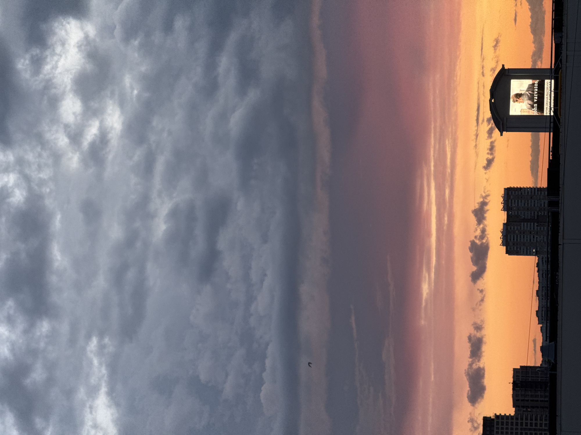 This is a vertical, eye-level shot of a cityscape at dusk, with a vibrant sunset filling the lower half of the frame. Heavy, dark grey clouds dominate the upper portion, transitioning to hues of purple and pink as they meet the bright orange sky below. A small bird is visible in flight against the muted purple clouds. The cityscape is composed of several modern, multi-story buildings, silhouetted against the warm glow of the sunset. On the right side of the frame, a prominent billboard stands out. It displays a portrait of a man wearing a jacket and the text "john varvatos." Below this, smaller text, partially obscured by glare, is visible. The billboard appears to be part of a structure with a white, arched roof. The foreground shows a flat rooftop or platform. The scene is devoid of any visible people, and the overall atmosphere is one of urban tranquility as day transitions into night. The location is identified as Sovkhoza imeni Lenina, Russia, suggesting a suburban or semi-urban area on the outskirts of a larger city.