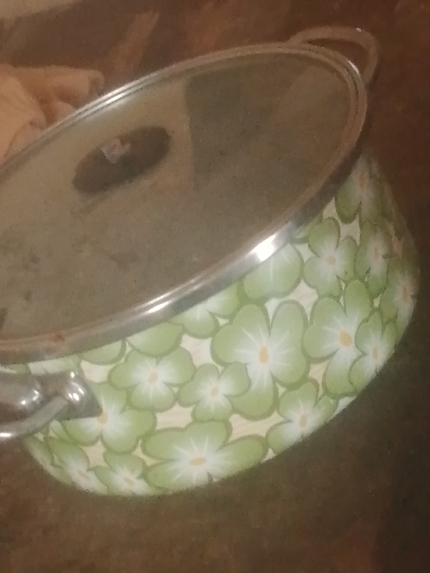 This media file is a close-up, slightly blurry, indoor shot of a patterned pot with a glass lid. The pot has a light-colored background adorned with light green flowers featuring white petals and yellow centers. The pot has a silver rim and a silver handle visible on the left side. The glass lid is reflective, showing a faint image of what appears to be a person's hand holding something, possibly a cloth, on the left side. The background of the scene is out of focus and appears to be a dark, textured surface, possibly a table or countertop, with hints of warmer tones. The lighting suggests an indoor setting, and there is no indication of the time of day or weather. The overall impression is a domestic scene, likely in a kitchen or dining area. There is no visible text in the image. The location context provided is Jauro Yinu, Nigeria.