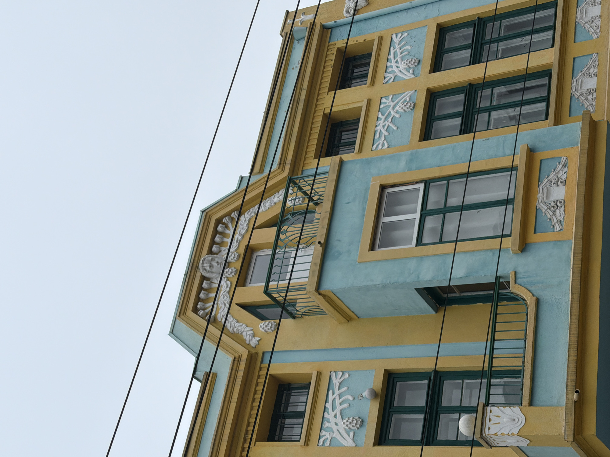 The image depicts the facade of a building in Varna, Bulgaria, characterized by a distinct architectural style featuring a color palette of yellow and light blue. The building's exterior is adorned with white, ornate relief patterns around the windows and along the edges. The windows themselves are primarily dark-framed, multi-paned units. A series of dark cables or wires extend across the foreground, partially obscuring the view of the building. A small, grilled balcony and a metal ladder are visible on the building's facade. The background is plain and light in color, likely the sky.
