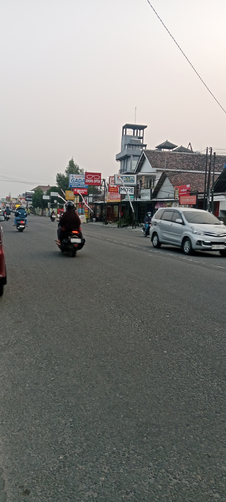 The image captures a street scene in Maguwoharjo, Indonesia, likely during late afternoon or early morning given the overcast sky. The road, paved with asphalt, shows signs of wear and tear with visible aggregate. Several motorbikes are on the road; one in the foreground has a rider wearing a dark helmet and jacket, and another motorbikes with riders is visible further down the road. A silver car is in the right lane, moving away from the camera.
The left side of the road is lined with buildings housing various businesses, indicated by numerous signs. Notable signs include "GADA EROPA OPTIK," "DAM," and "MORE," suggesting a commercial area. Some buildings have a traditional architectural style with tiled roofs, while others appear more modern. A prominent multi-story structure with a tower-like feature stands on the right side of the street, dominating the skyline.
The overall atmosphere is busy, with ongoing traffic and commercial activity. The sky is a uniform pale grey, indicating diffused light conditions. Trees are visible behind some of the buildings on the left.