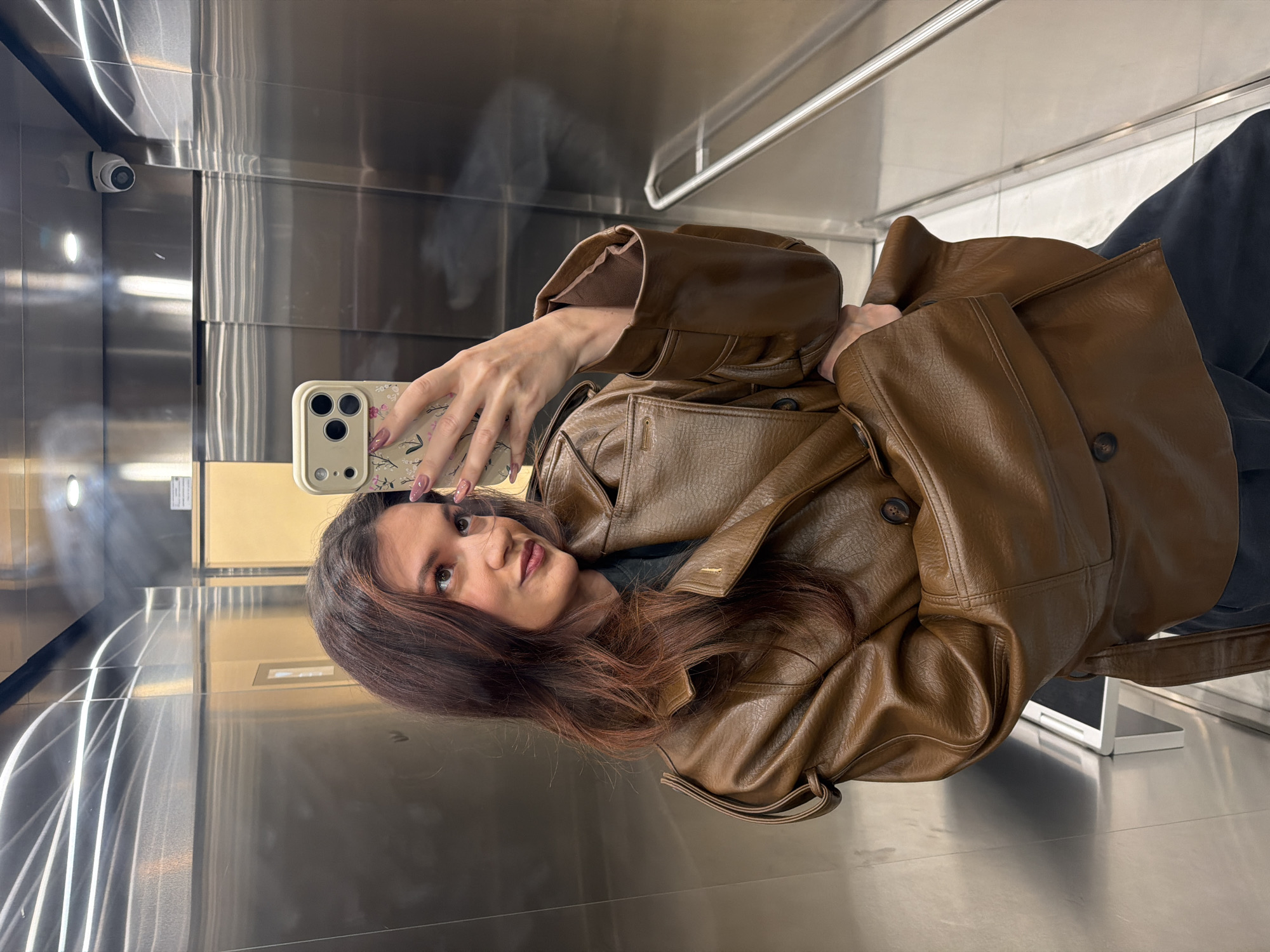 A young woman with shoulder-length brown hair is taking a selfie in what appears to be an elevator with reflective metal walls. She is wearing a brown leather jacket and holding a smartphone with a floral patterned case. A security camera is visible in the upper left corner. The scene could be in any modern building in Belgrade, Serbia, as elevators are common in many structures and the details don't offer specific location clues.