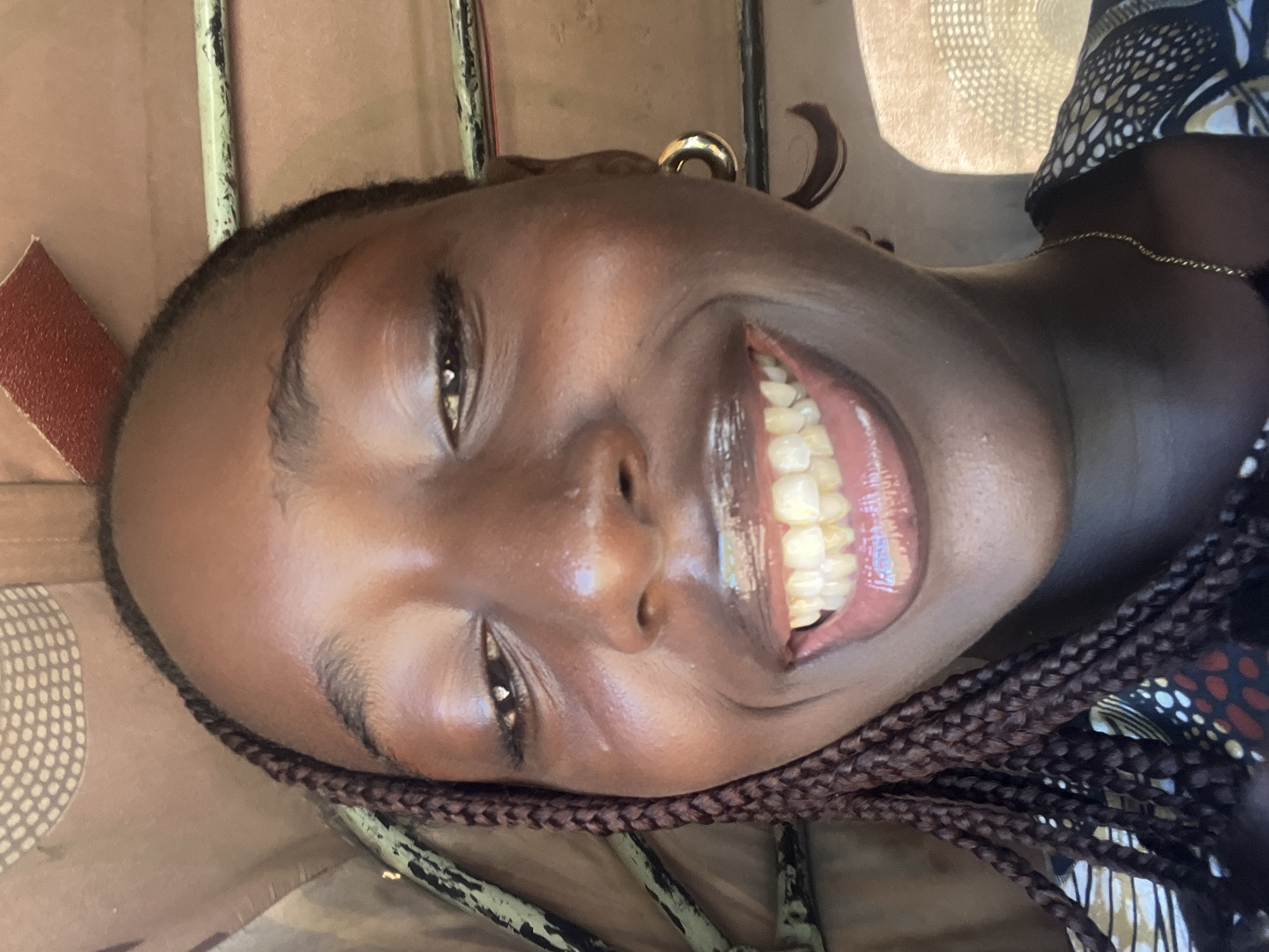 A close-up portrait shot captures a young individual, likely female, of African descent, smiling broadly directly at the camera. Her dark skin exhibits highlights across the forehead, nose, cheeks, and chin, suggesting a glossy complexion. Dark, braided hair is visible on the left side of her face. Her eyes, appearing dark, are partially closed due to the wide smile, with distinct light reflections in the irises. Her lips are coated with a light-colored gloss, revealing both upper and lower teeth in the open-mouth smile. A single gold-colored hoop earring is present on the right ear, and a thin gold-colored chain necklace is visible around the neck. The subject is wearing a garment with a complex pattern of white, black, and reddish-brown shapes. The background indicates an interior setting, possibly within a vehicle or small structure in Galadimawa Village, Nigeria. Visible elements include a light brown, possibly fabric-lined ceiling with a faint dotted pattern, and multiple greenish-grey metallic bars or pipes running horizontally and diagonally across the upper and left portions of the frame. A small section of reddish, textured material is discernible in the upper right corner.