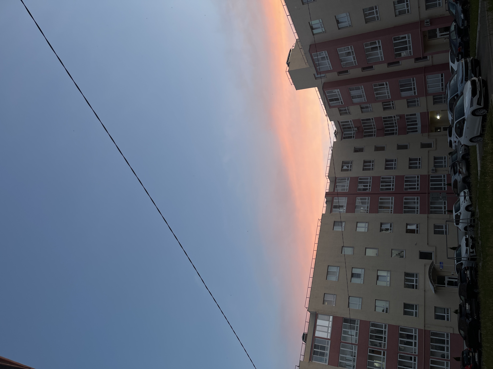 The image, rotated, depicts a multi-story residential building against a vibrant sky during either sunrise or sunset in Chaikovsky, Russia. The building, occupying the right side of the frame, is modern with alternating sections of light beige and reddish-pink on its facade. Numerous rectangular windows with white frames and some balconies with dark railings are visible across its many floors.
To the immediate right of the building, a parking area or street is visible with several cars parked in a row. These vehicles include various models and colors, such as dark, white, and grey. A narrow strip of green grass separates the building from the parked cars. Faint Cyrillic characters are visible on some car license plates, consistent with the Russian location.
The sky, dominating the left and upper portions of the image, presents a striking color gradient. It transitions from a deep blue on the far left to soft pink, orange, and light blue hues closer to the horizon line where it meets the building. This warm coloration strongly suggests either dawn or dusk. A single, dark, thin cable or power line stretches diagonally across the sky from the top-left to the bottom-right of the frame.
No people are visible, and no specific activities are taking place. The overall scene captures a serene moment in an urban or suburban environment, characterized by the dramatic sky colors and modern architecture.